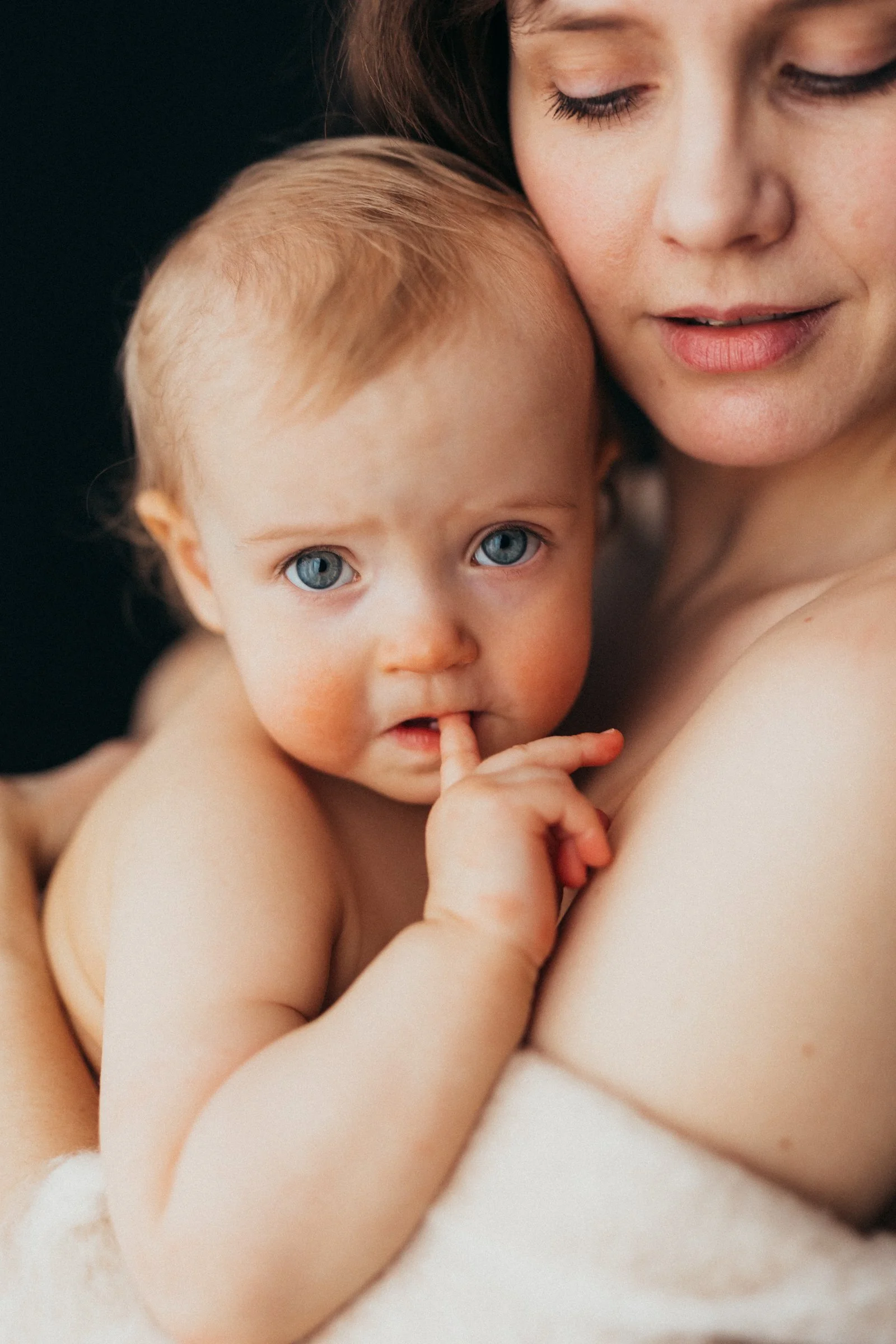 A woman holding a baby with blue eyes and light skin, the woman has closed eyes and a gentle expression, both appear to be cuddling against a dark background.