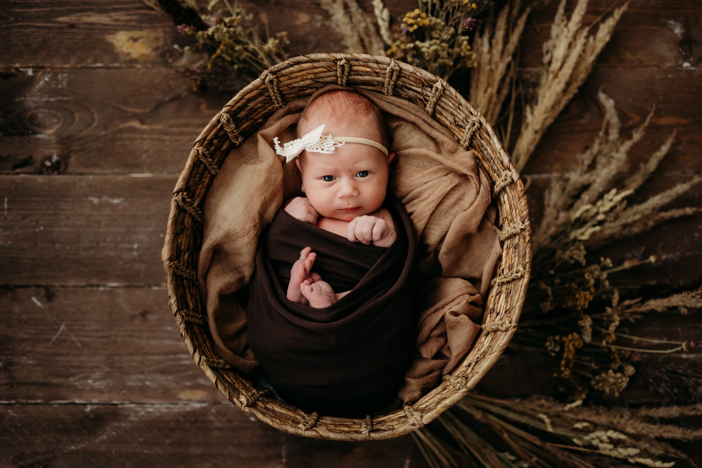 A newborn baby girl lying in a woven basket, wrapped in a dark brown blanket and surrounded by beige and brown soft fabrics, with a cream-colored lace headband with a bow on her head, on a wooden floor with dried flowers scattered around.