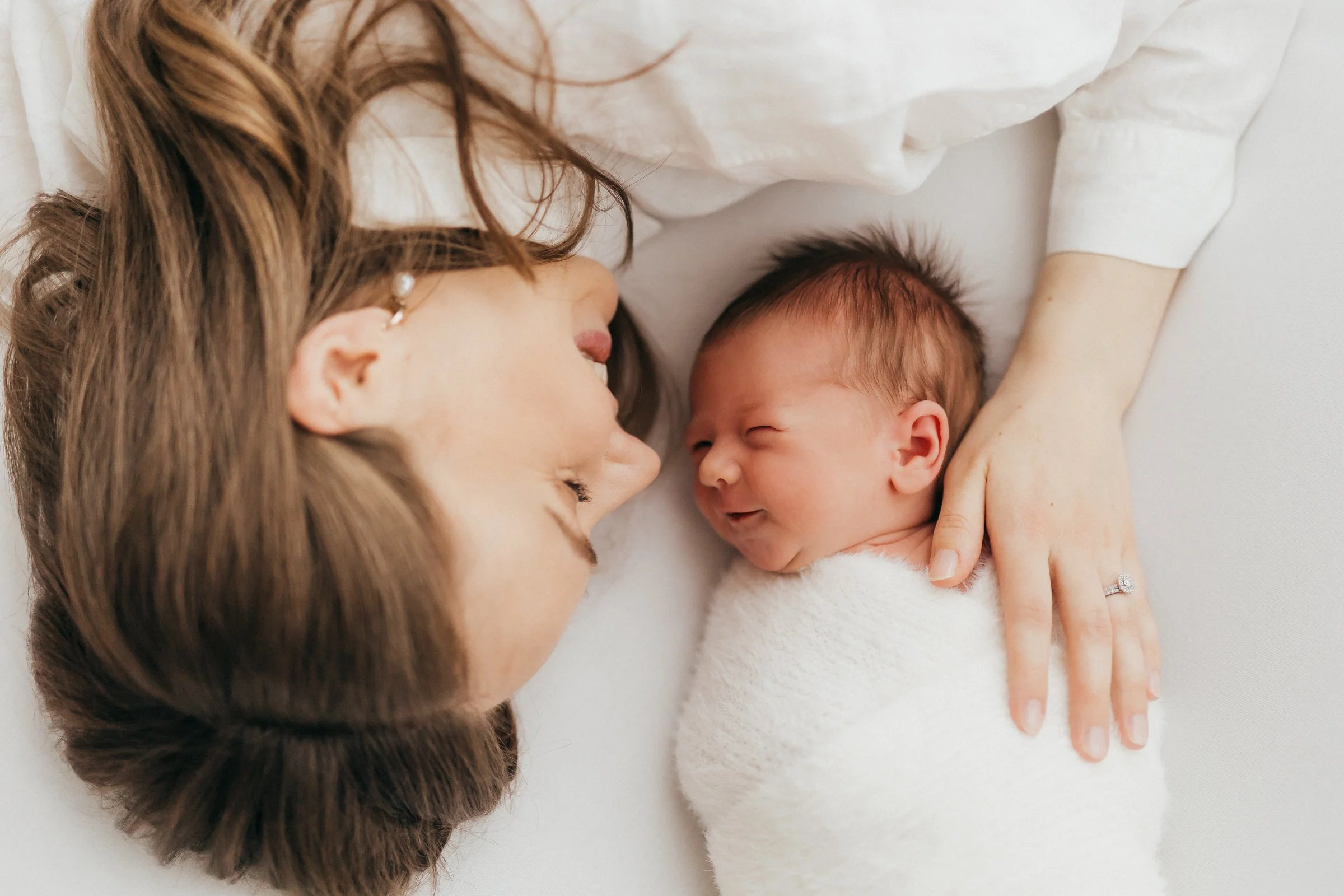 A woman with brown hair and pearl earrings lying next to a newborn baby wrapped in a white blanket, both smiling and touching foreheads.
