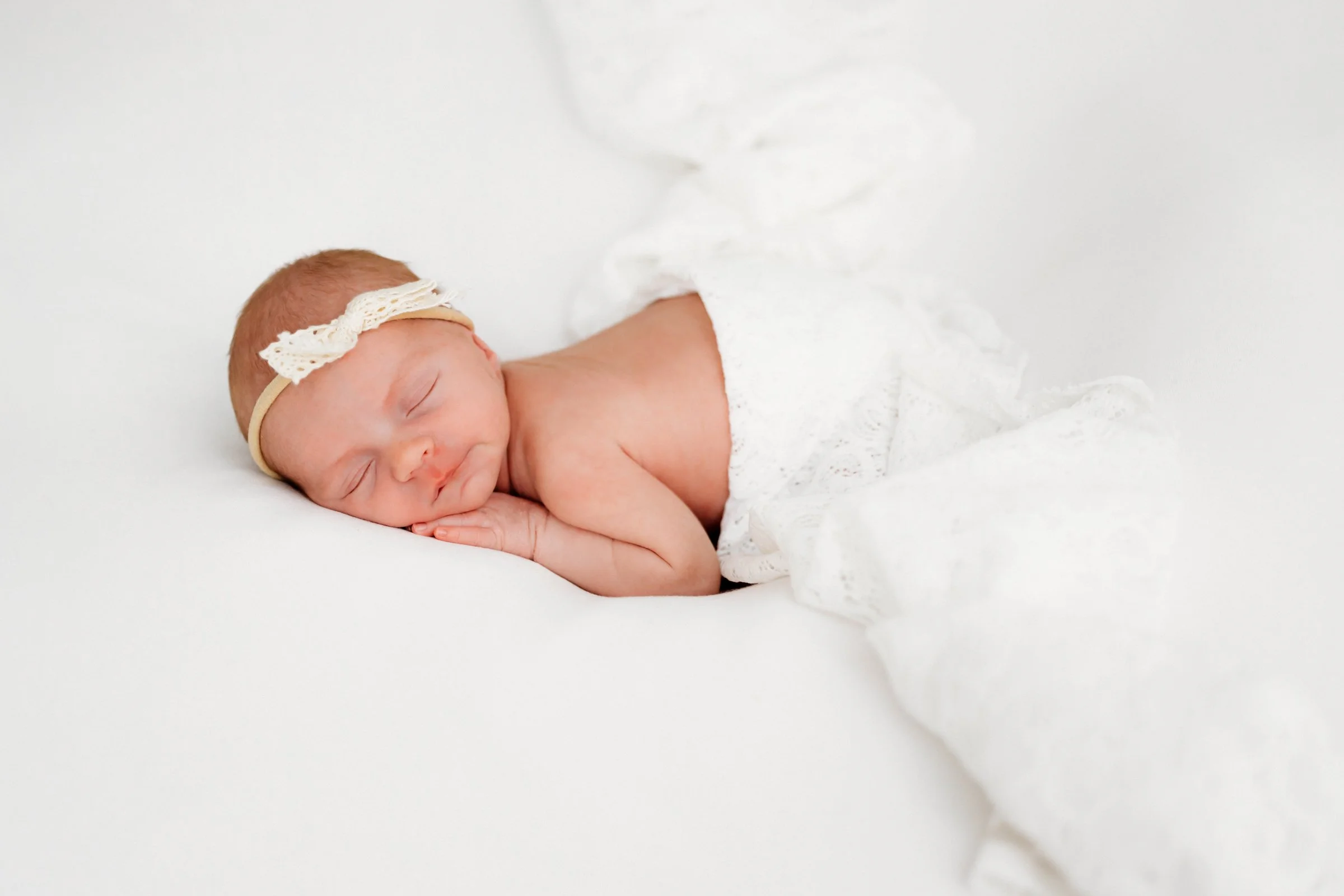 Baby sleeping on a white surface, wearing a lace headband and partially covered with a white blanket.
