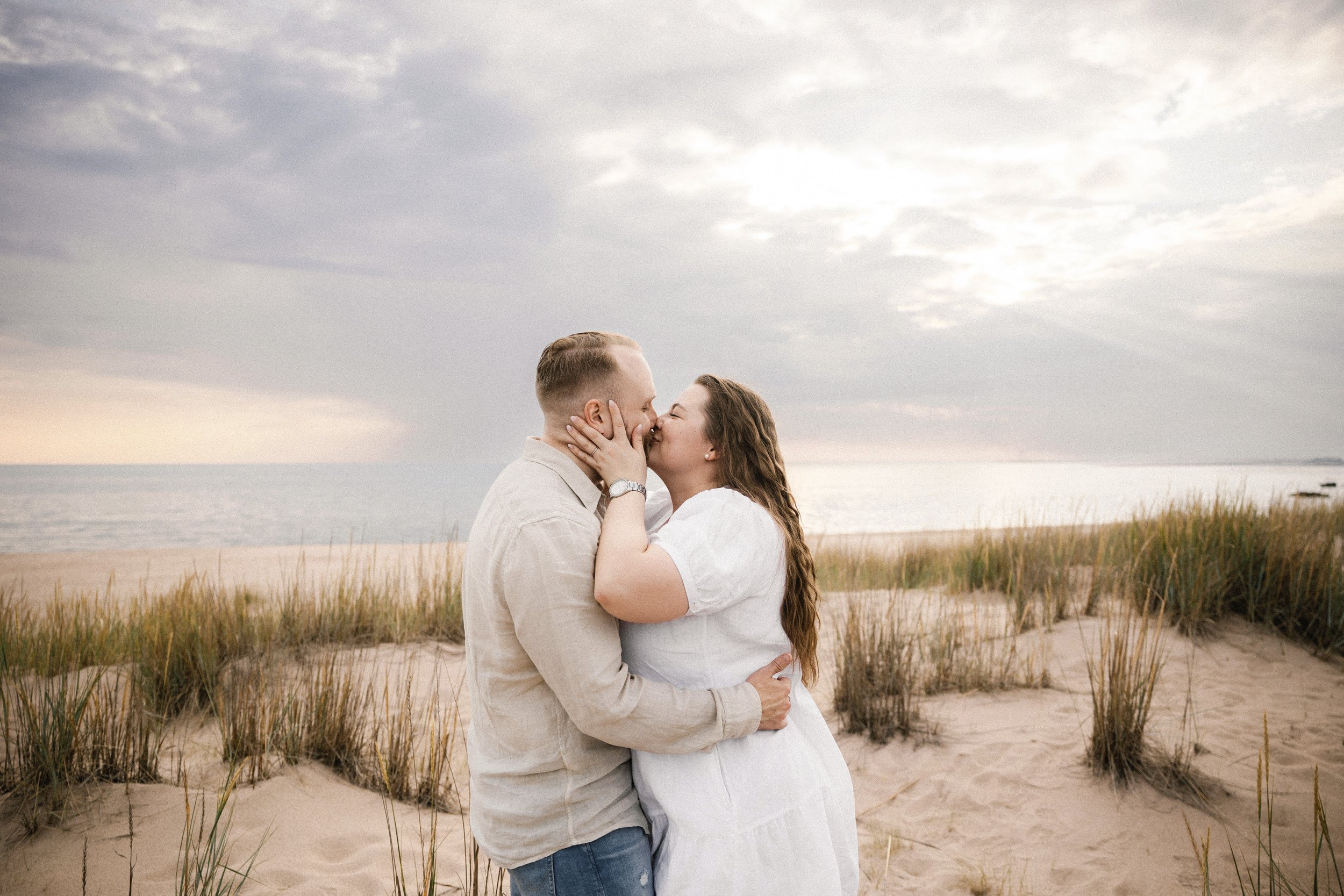 A couple kissing on a sandy beach with grasses, the ocean, and a cloudy sky in the background.
