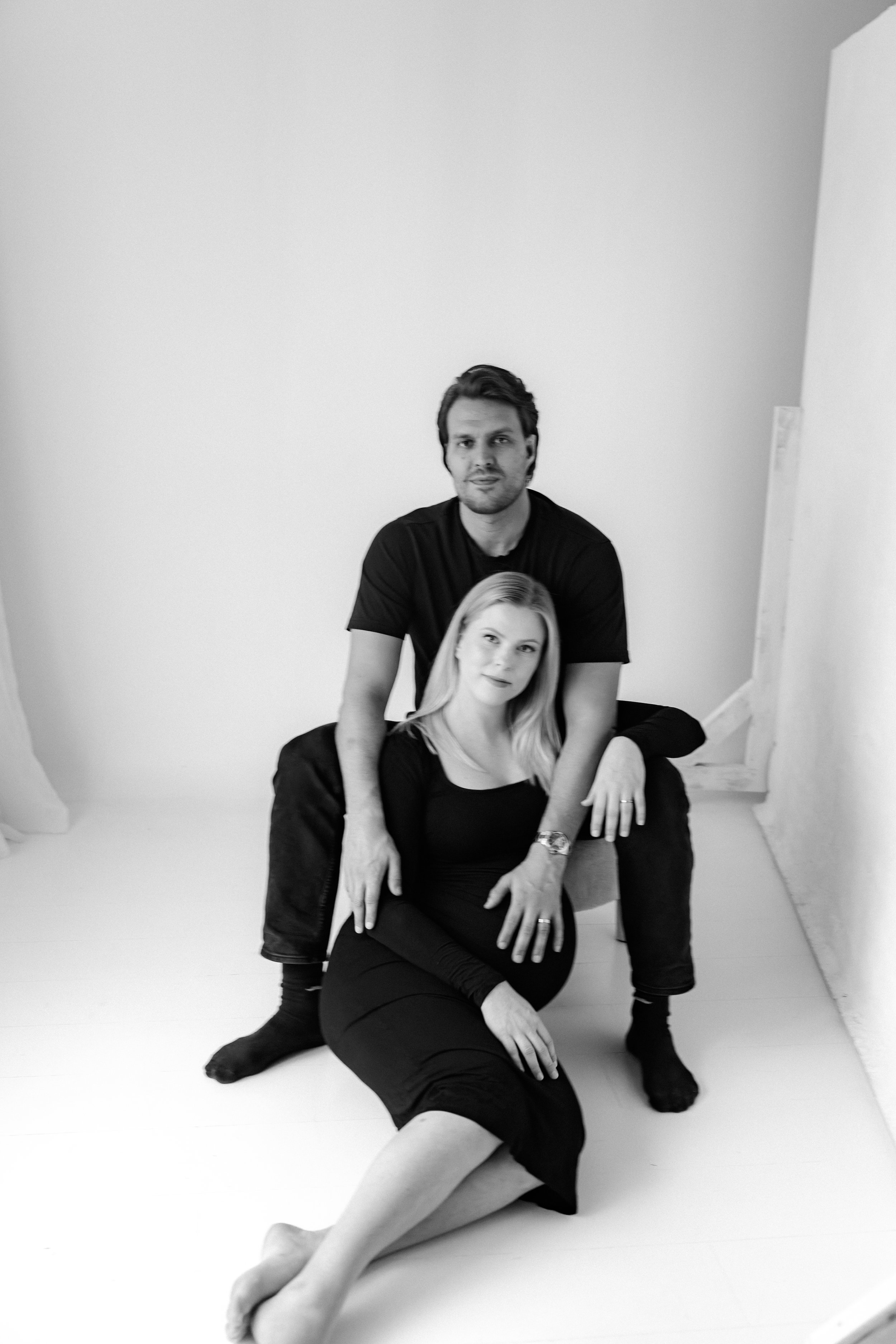 A black and white photo of a man and woman posing together in a studio with plain white walls. The woman is sitting on the floor with her legs extended and her hand resting on her knee, while the man is seated behind her, with one hand on her shoulde