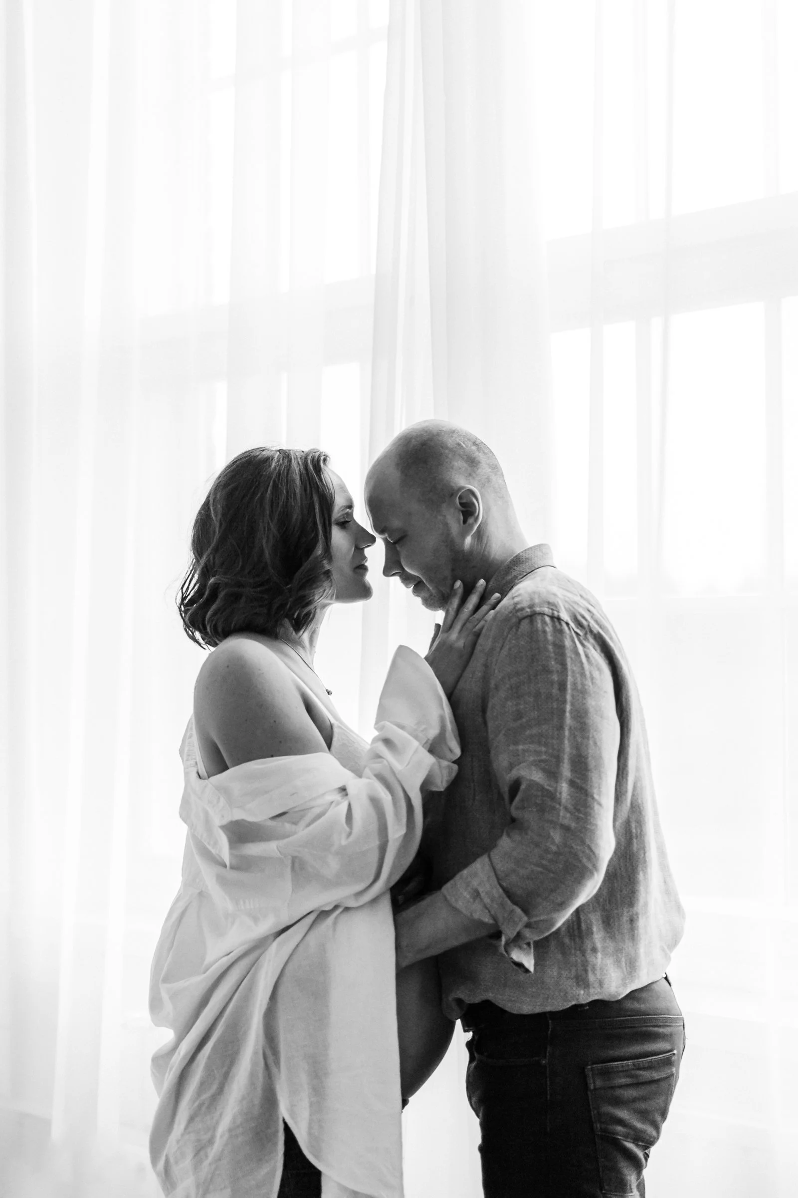 A black-and-white intimate photo of a couple, with the woman touching the man's face and both of their foreheads touching in front of a window with sheer curtains.