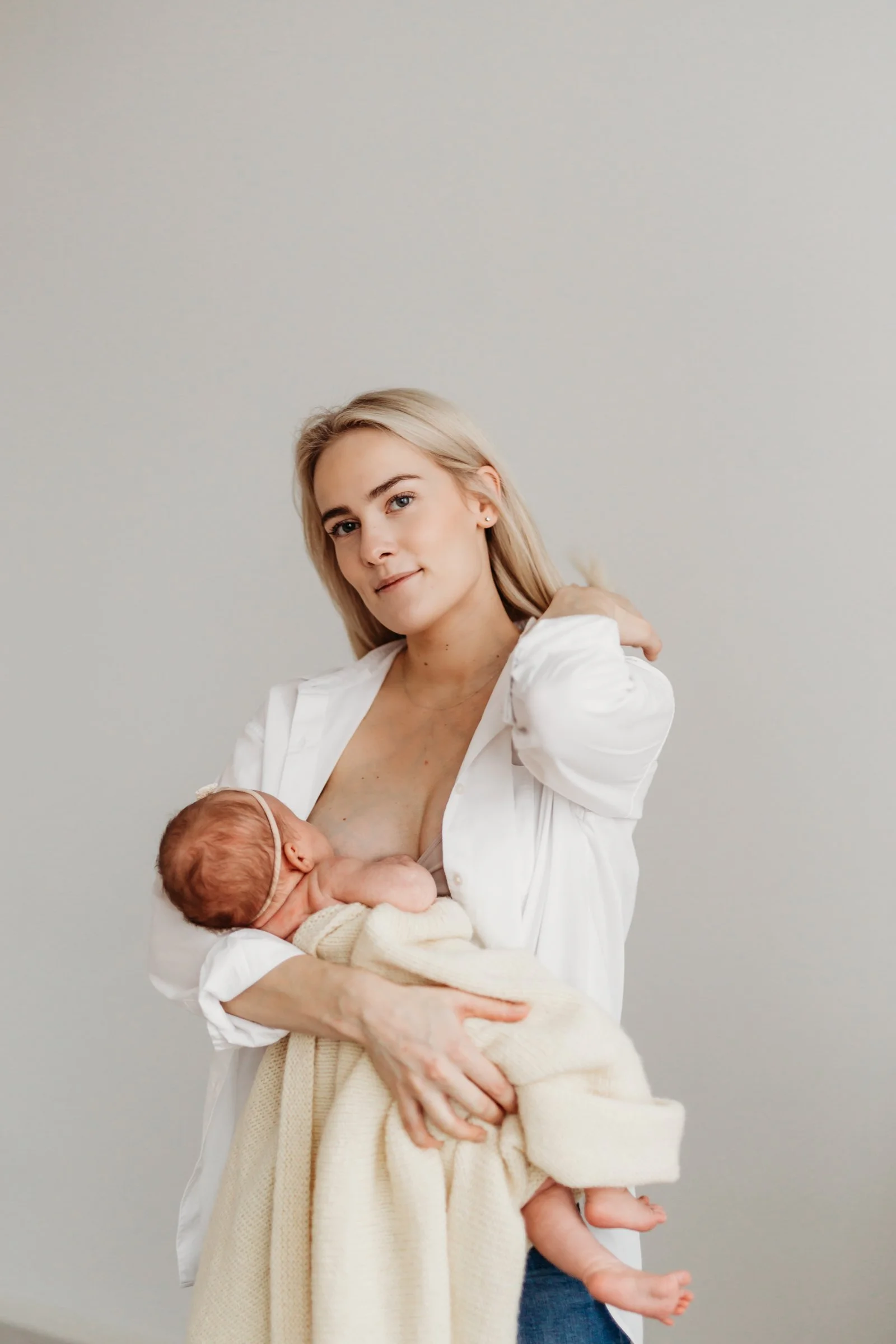 A woman with blonde hair breastfeeding a newborn baby wrapped in a cream blanket.