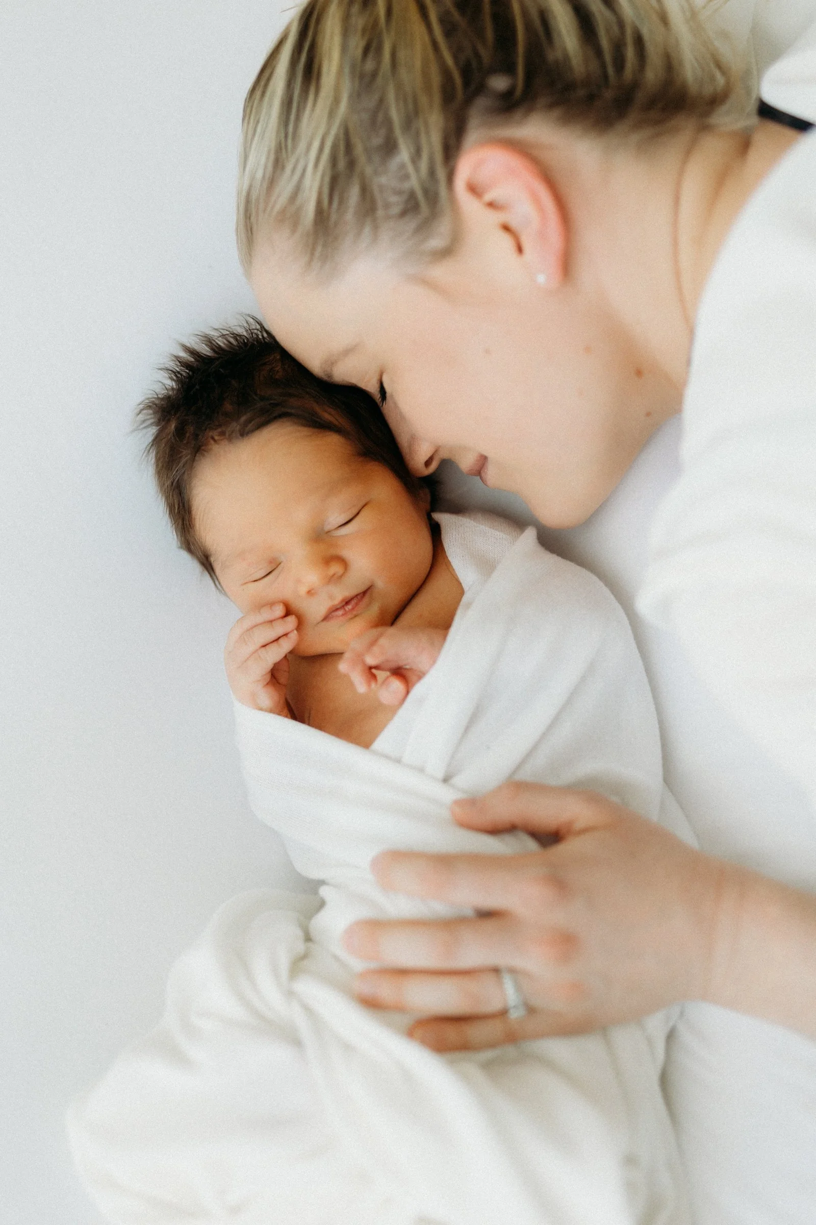 A mother holding her newborn baby wrapped in a white blanket, both with their foreheads touching and eyes closed, sharing a tender moment.