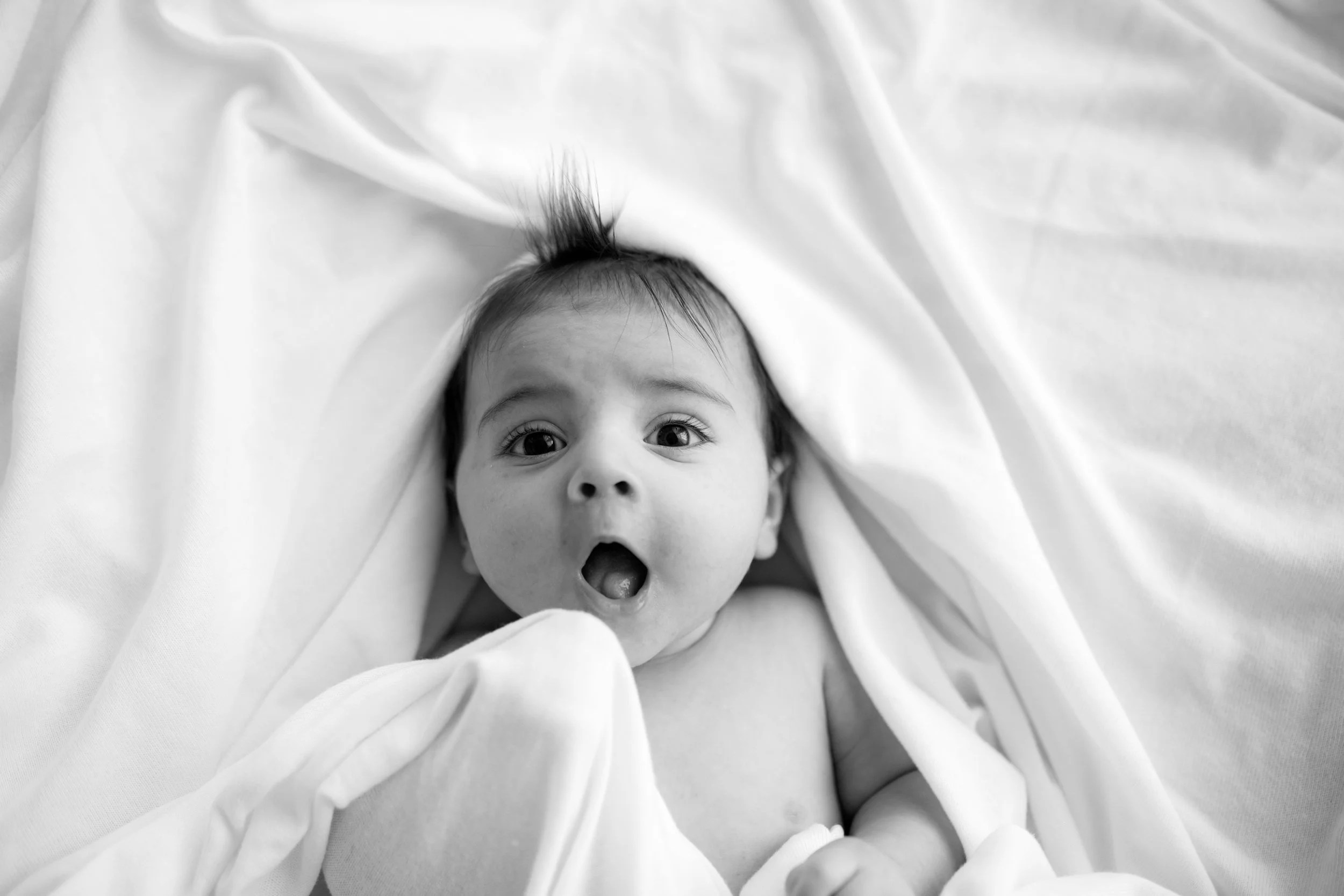 Black and white photo of a surprised baby lying on a soft surface, partially covered with a blanket, with wide eyes and an open mouth.