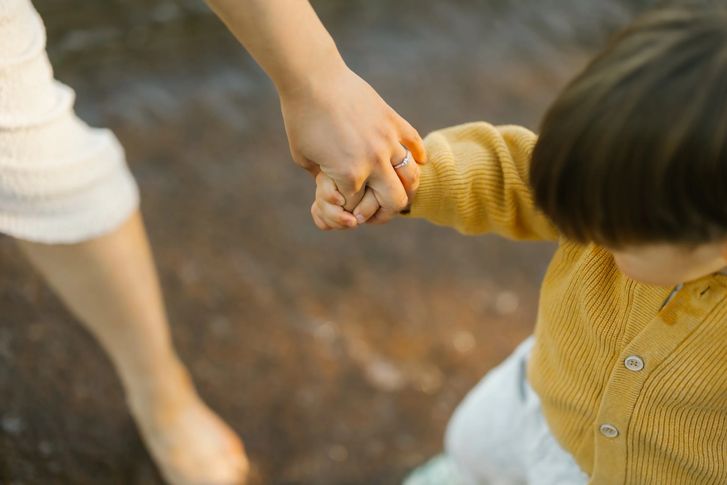 A woman holding a young boy's hand, guiding him as they walk on a dirt path outside.
