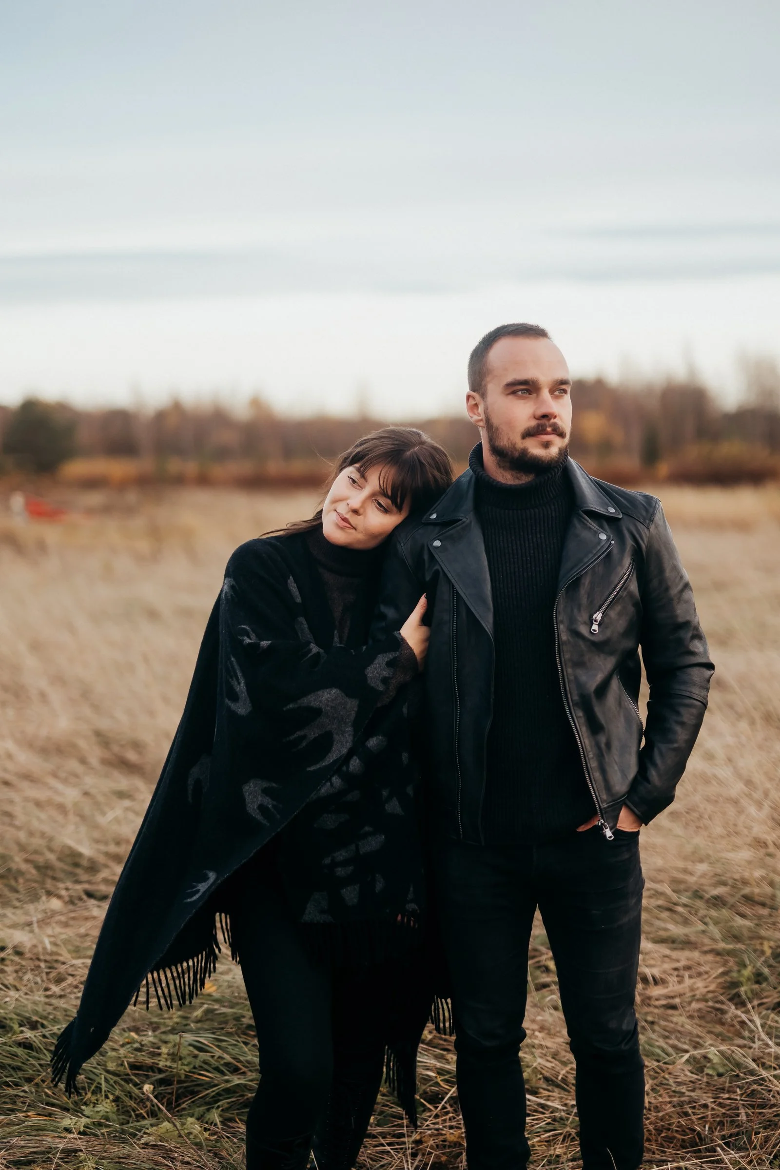 A man and a woman standing outdoors in a grassy field during autumn. The woman rests her head on the man's shoulder, and they both appear contemplative. The man wears a black leather jacket and black pants, while the woman wears a black patterned shawl or blanket.