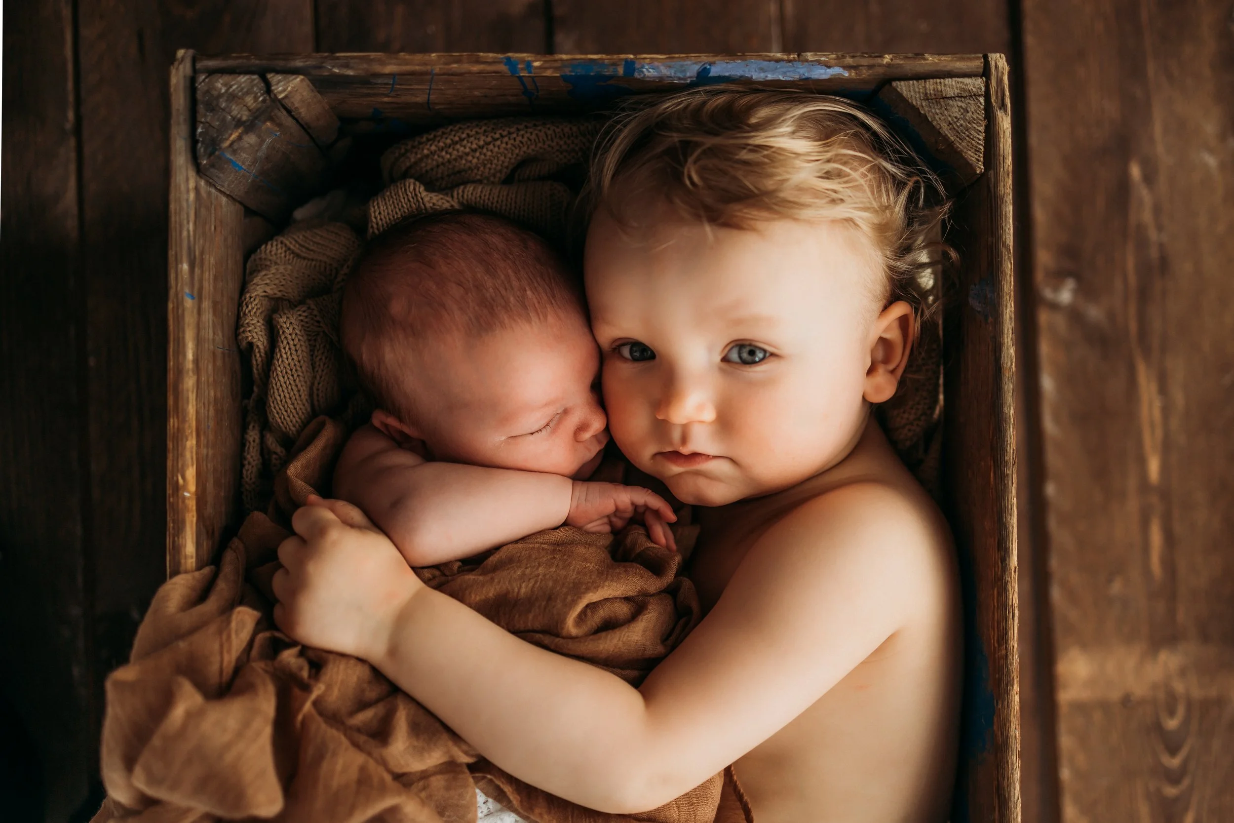 A young boy gently holding a newborn baby in a wooden box, both gazing at the camera. The baby is wrapped in brown cloth and the boy has curly blond hair, with a serious expression.