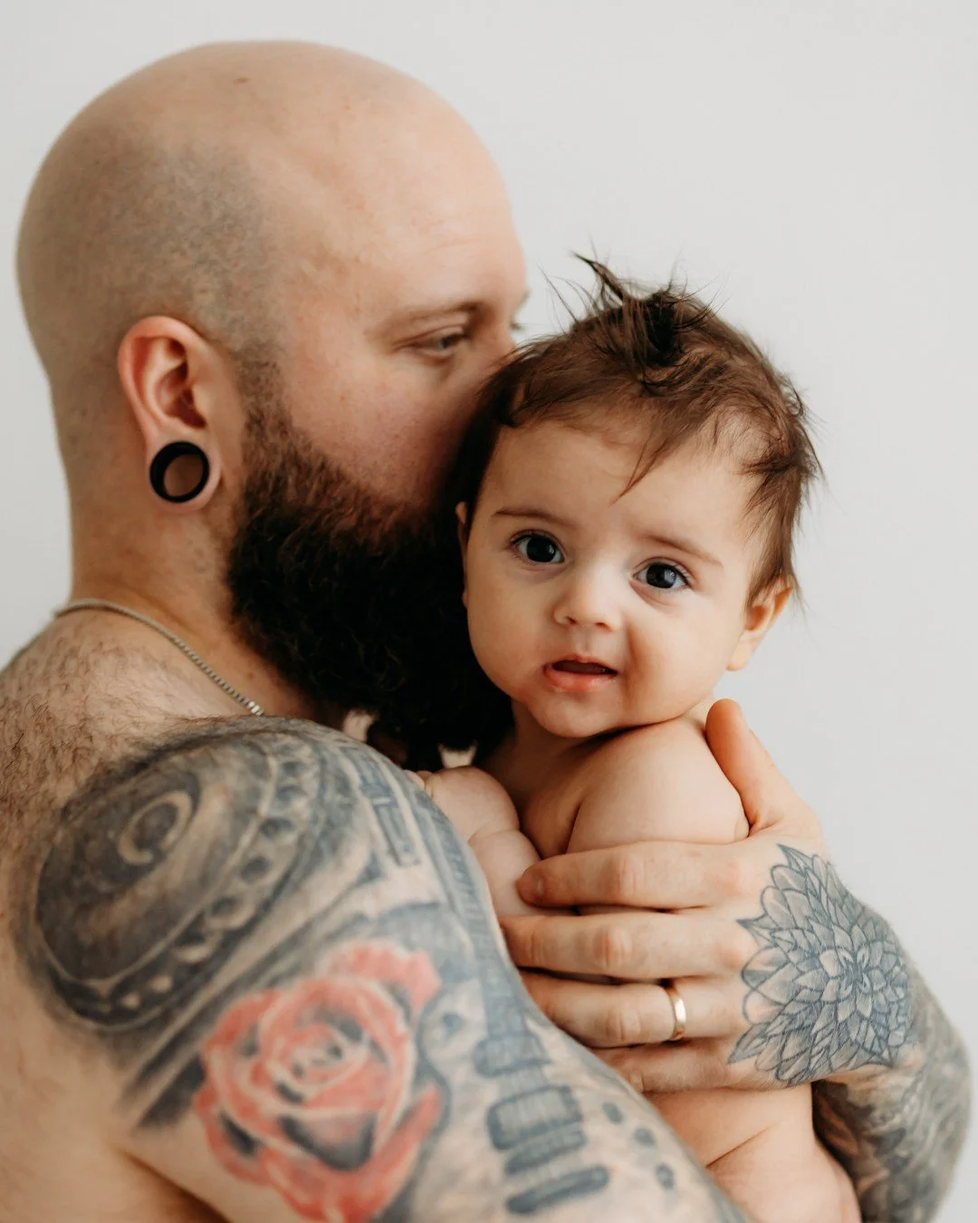 A shirtless man with tattoos, a beard, and earrings holds a young child with messy brown hair, blue eyes, and a curious expression, against a plain background.