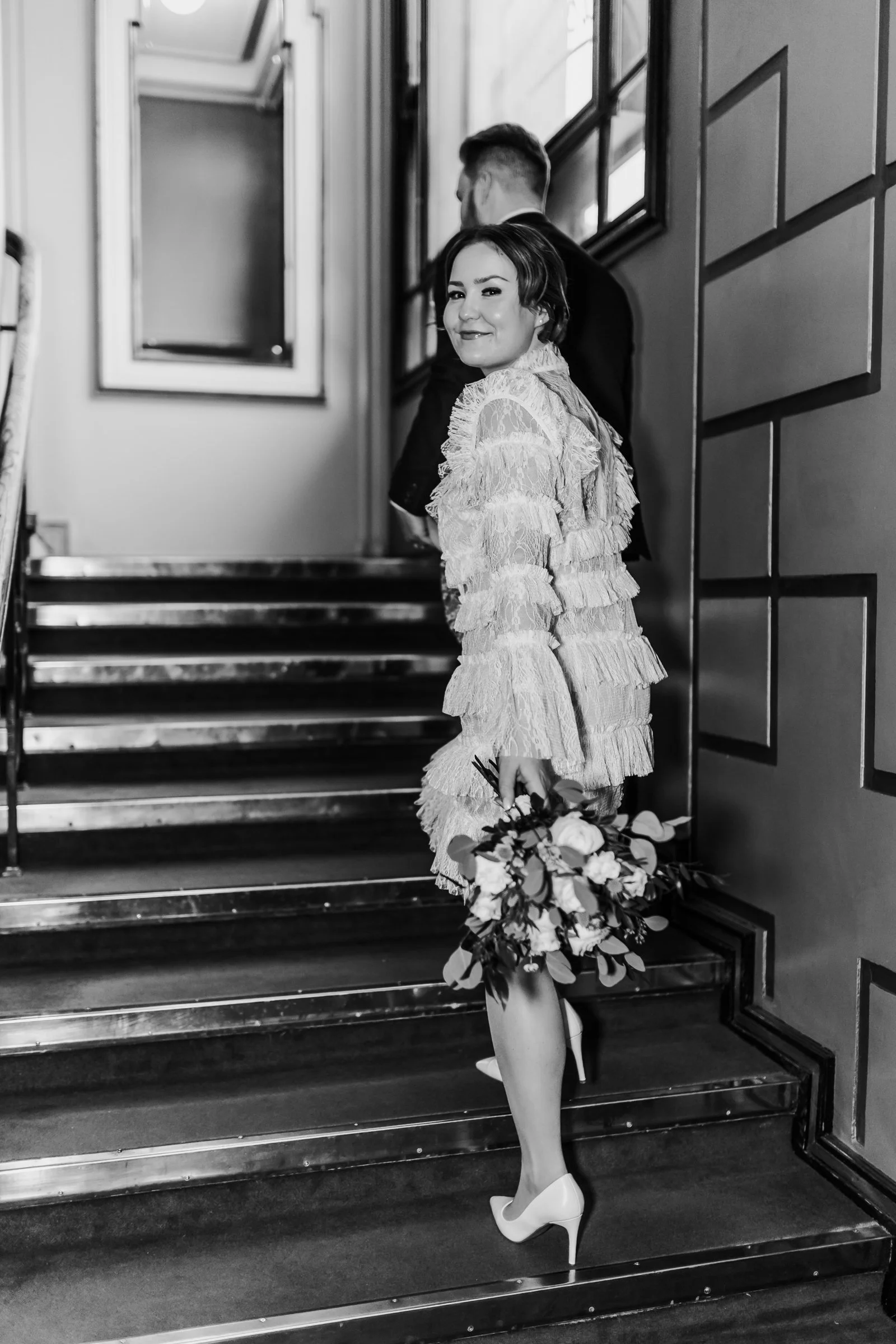 A woman in a lace dress holding a bouquet of flowers, standing on stairs inside a building, with a man in a suit behind her, both facing away from the camera.