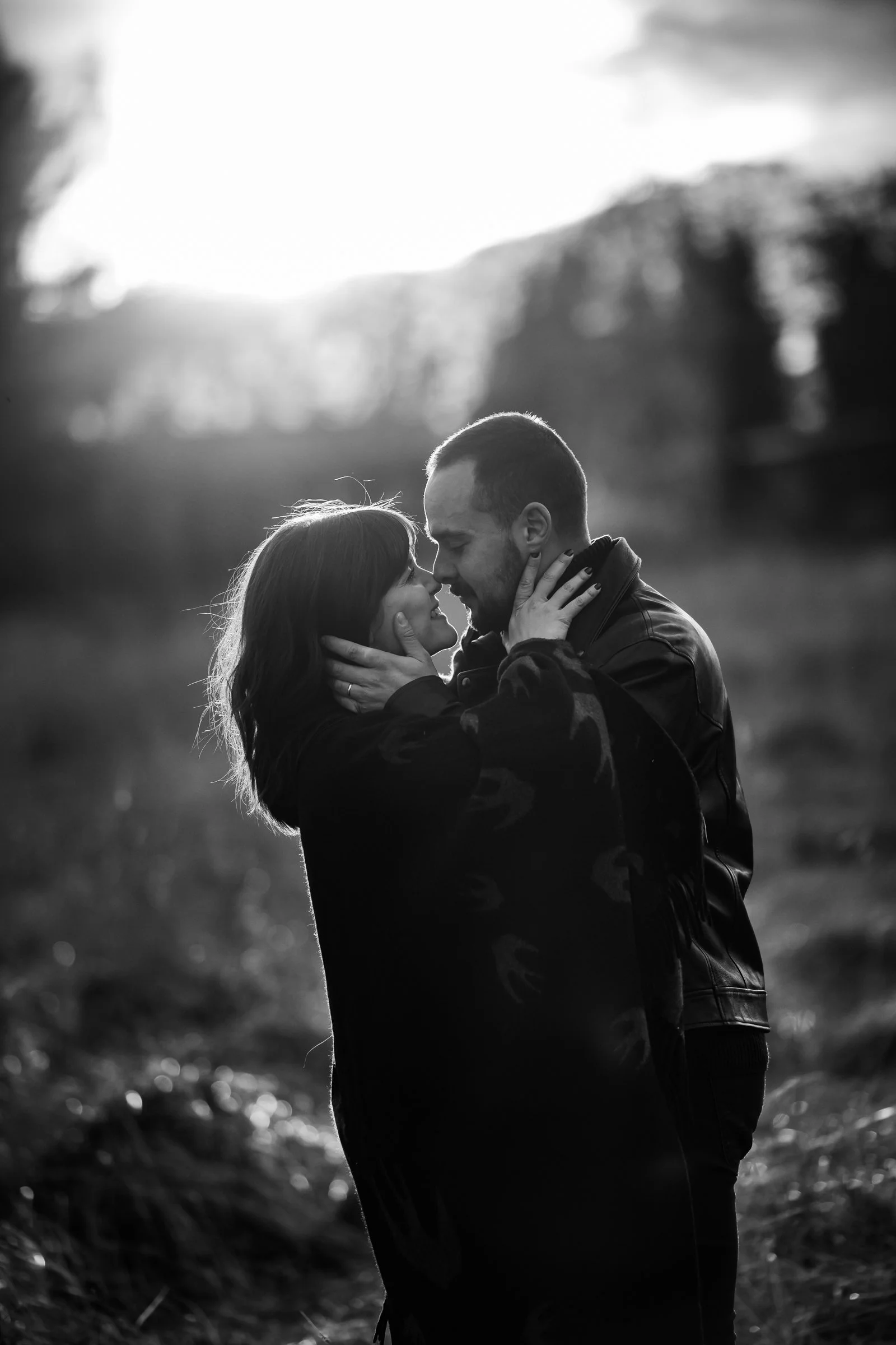 A black-and-white photo of a couple facing each other with close faces, touching noses, and embracing outdoors