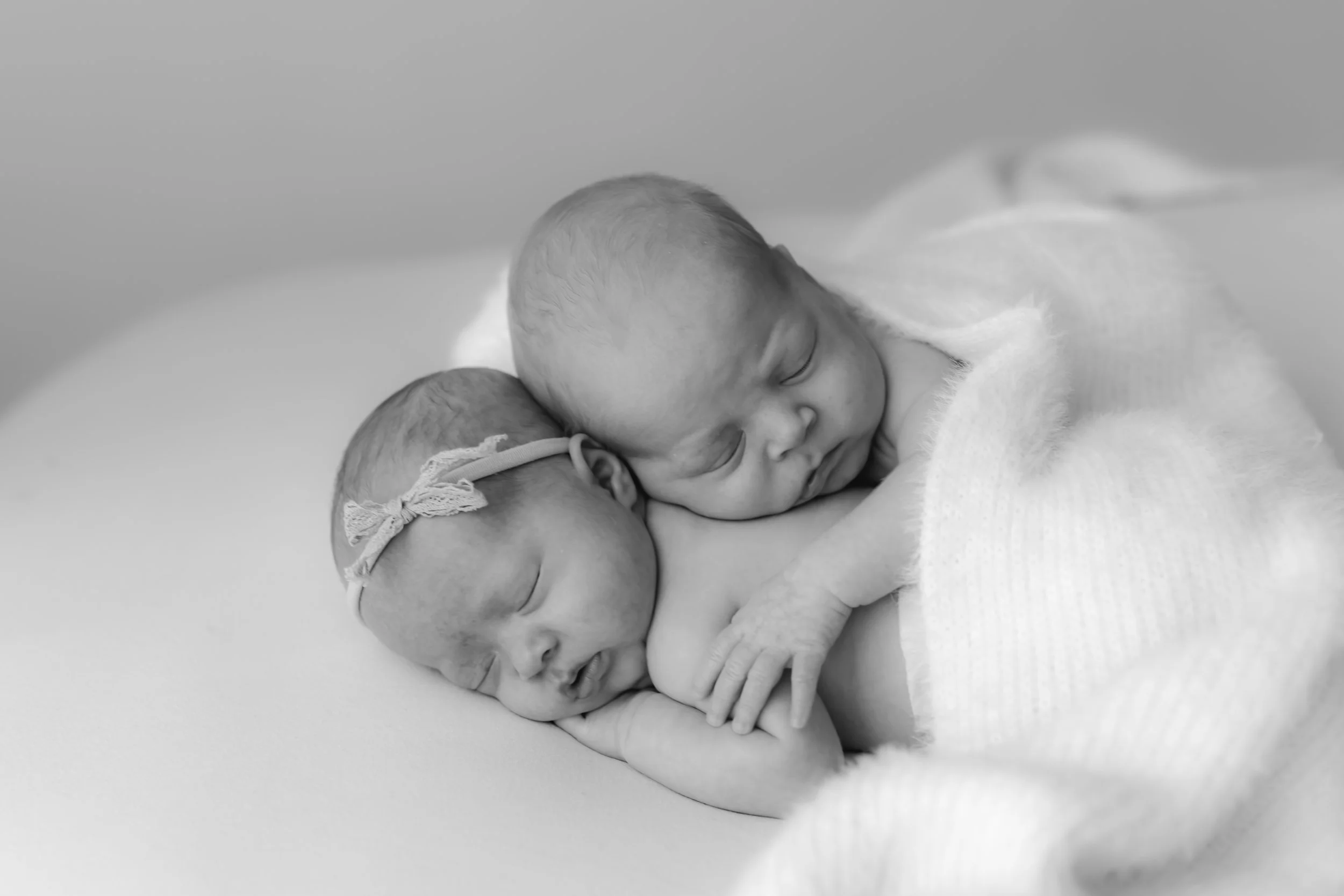Two newborn babies sleeping closely, one with a headband, wrapped in a soft blanket, in black and white.