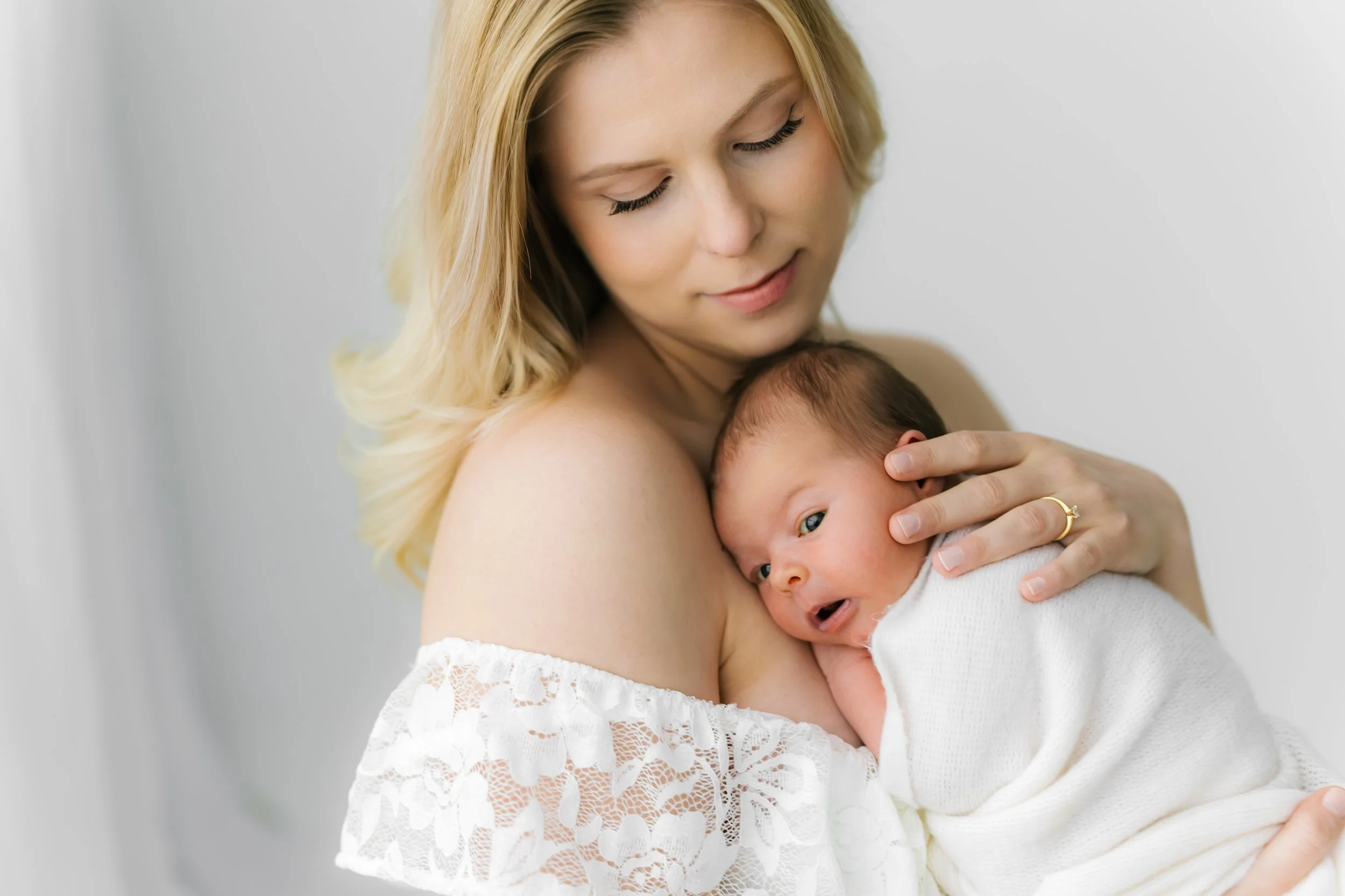 A woman with blonde hair holding a newborn baby wrapped in a white blanket, both looking affectionately at each other.
