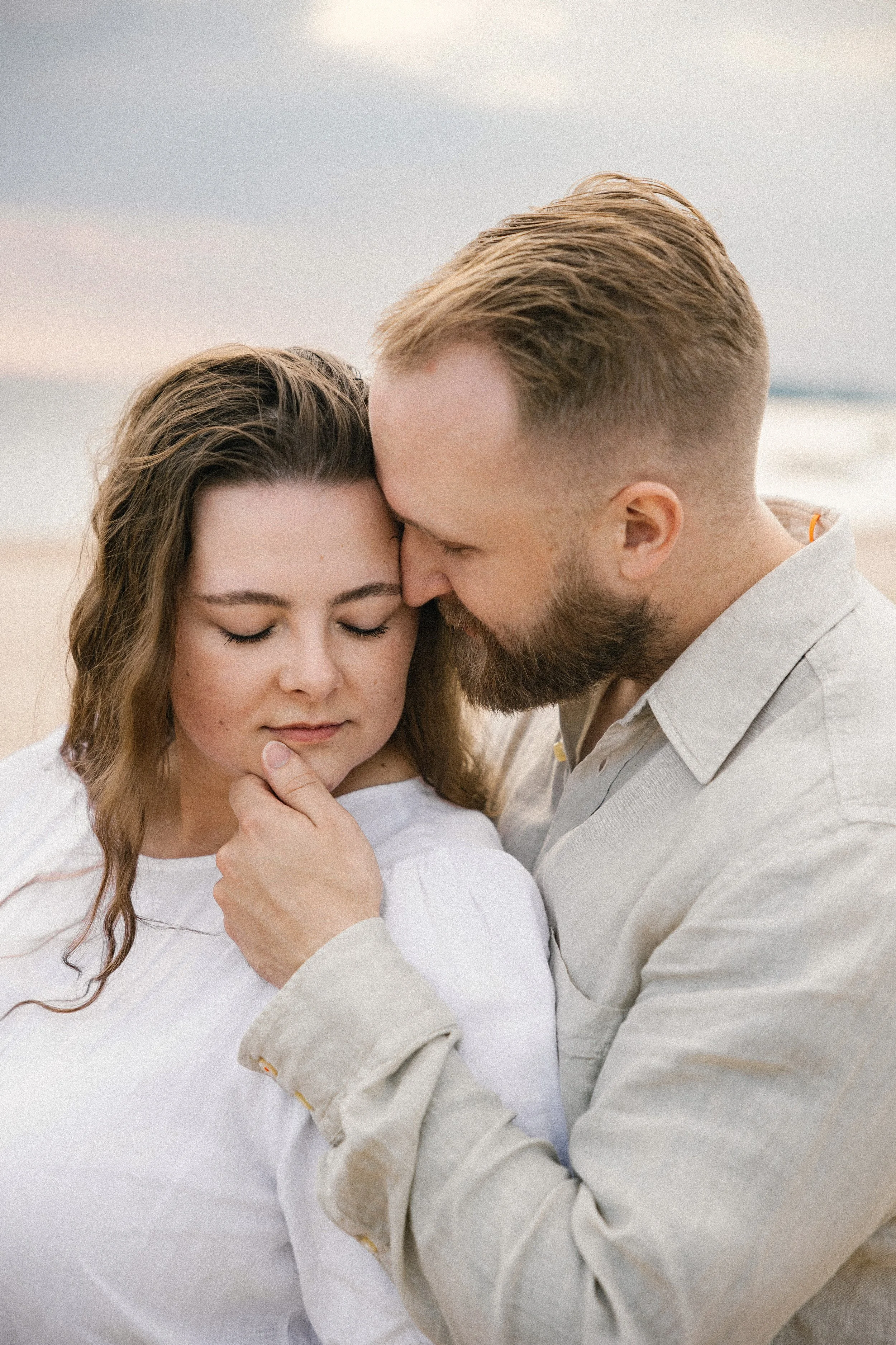 A romantic couple with their foreheads touching and eyes closed on a beach at sunset.