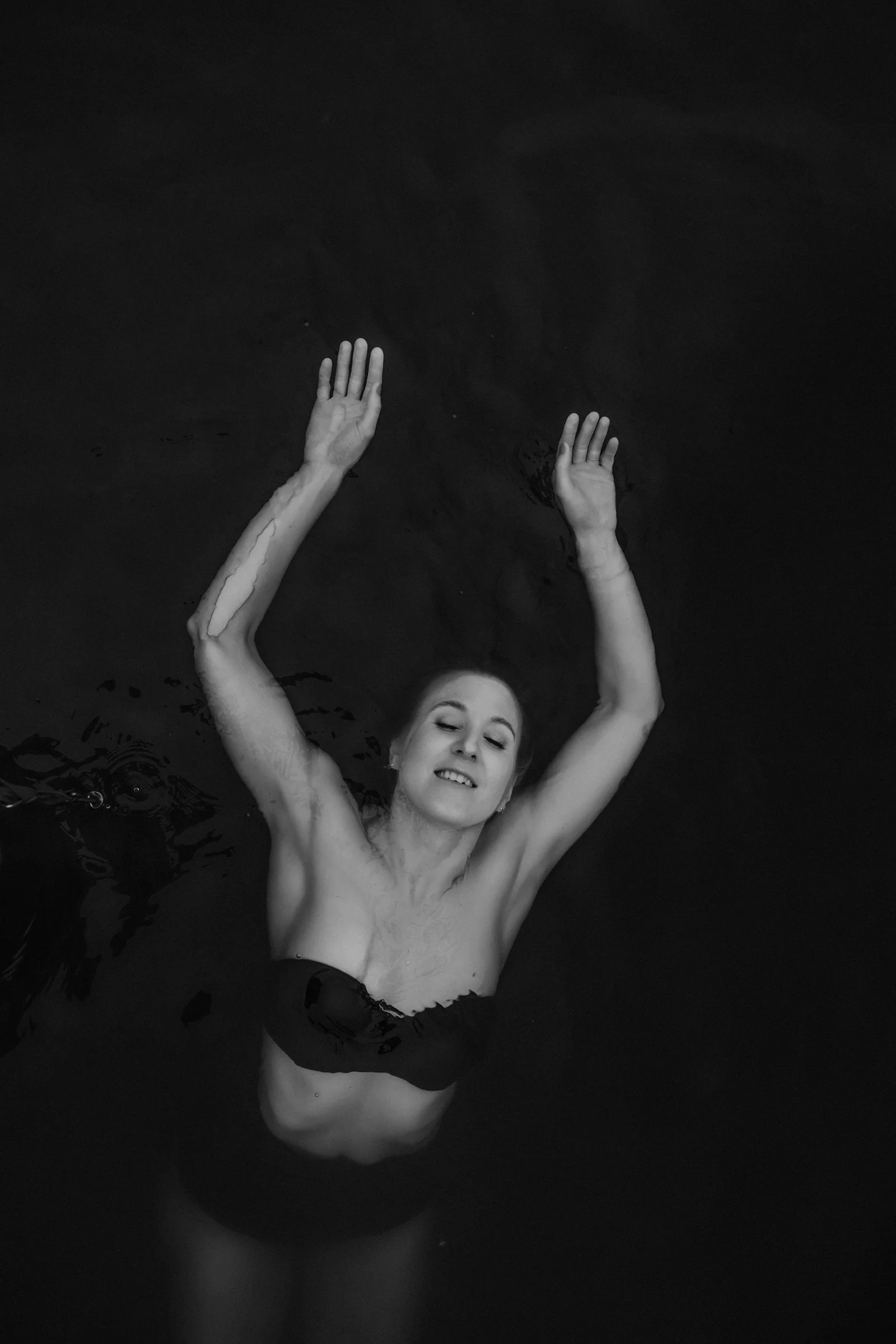 A woman swimming in dark water with arms raised, smiling with eyes closed.