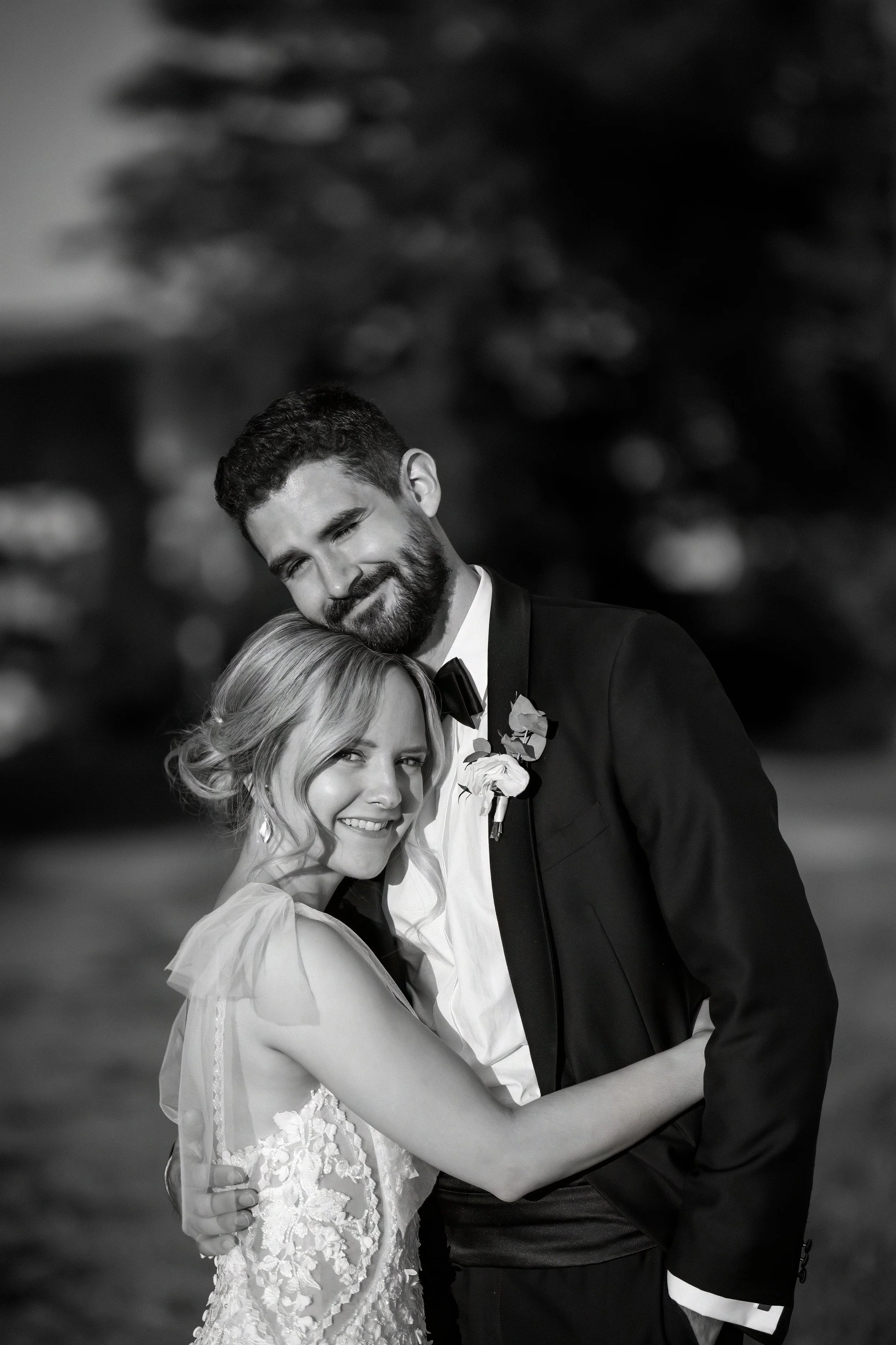 A black and white photo of a newlywed couple hugging outdoors at night, smiling, with blurred trees in the background.