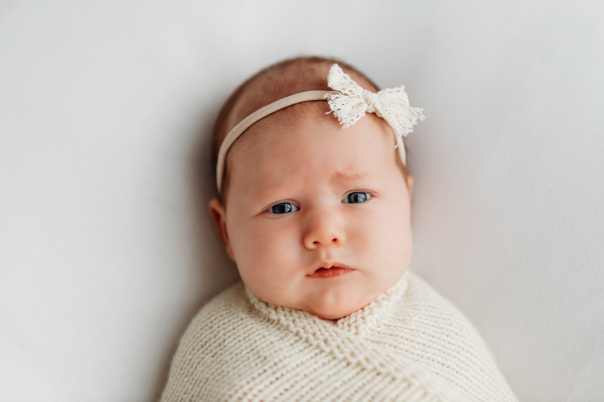 Close-up of a baby with blue eyes wearing a cream-colored knitted sweater and a lace headband with a bow, lying on a white surface.
