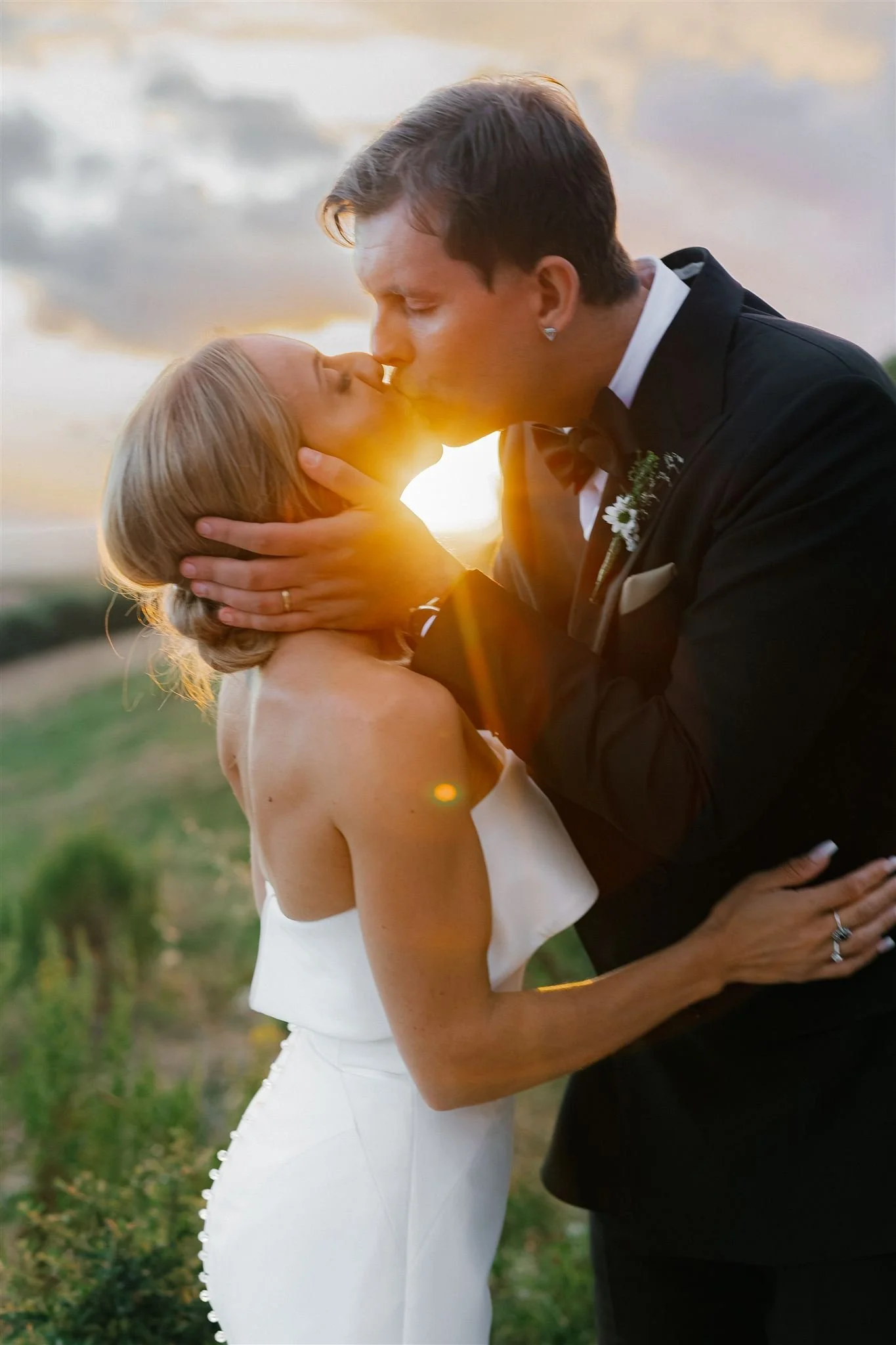 A bride and groom kissing outdoors during sunset, with the sun shining behind them.