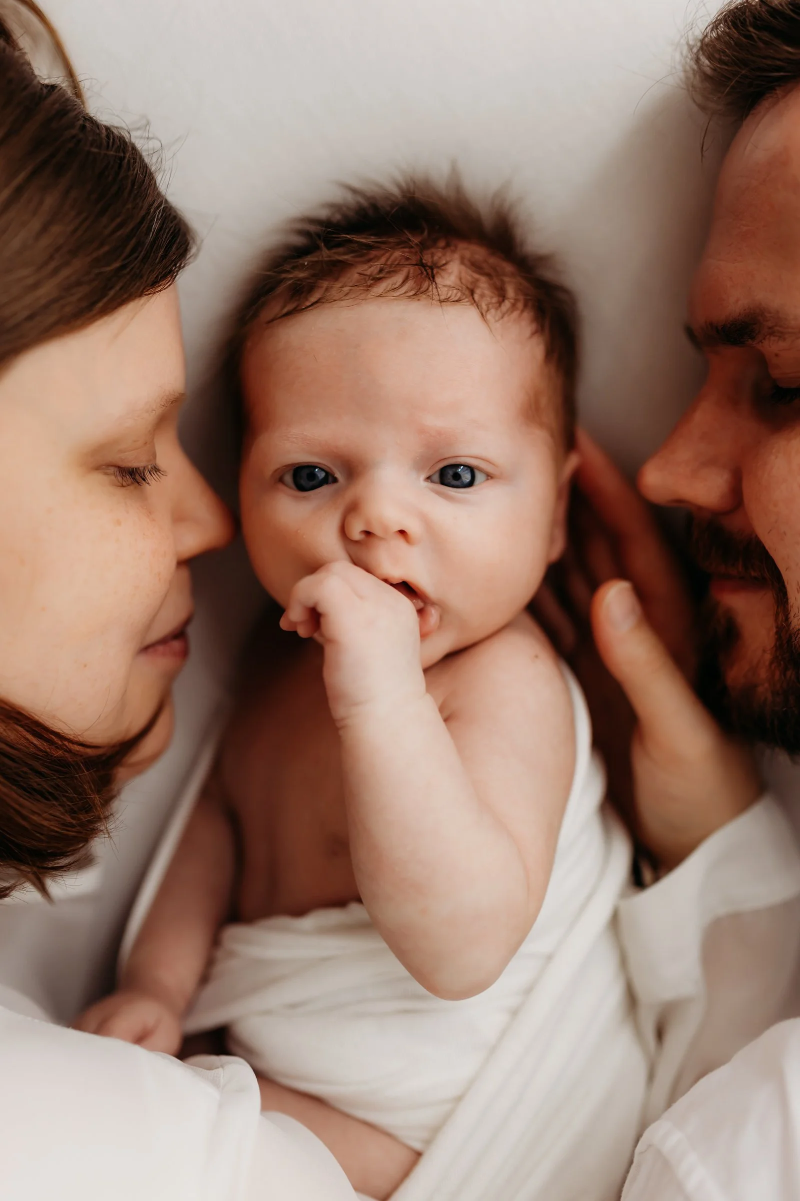 A baby with blue eyes lying between two adults, possibly the parents, who are gazing at the baby lovingly. The baby has its finger in its mouth and is wrapped in a white blanket.