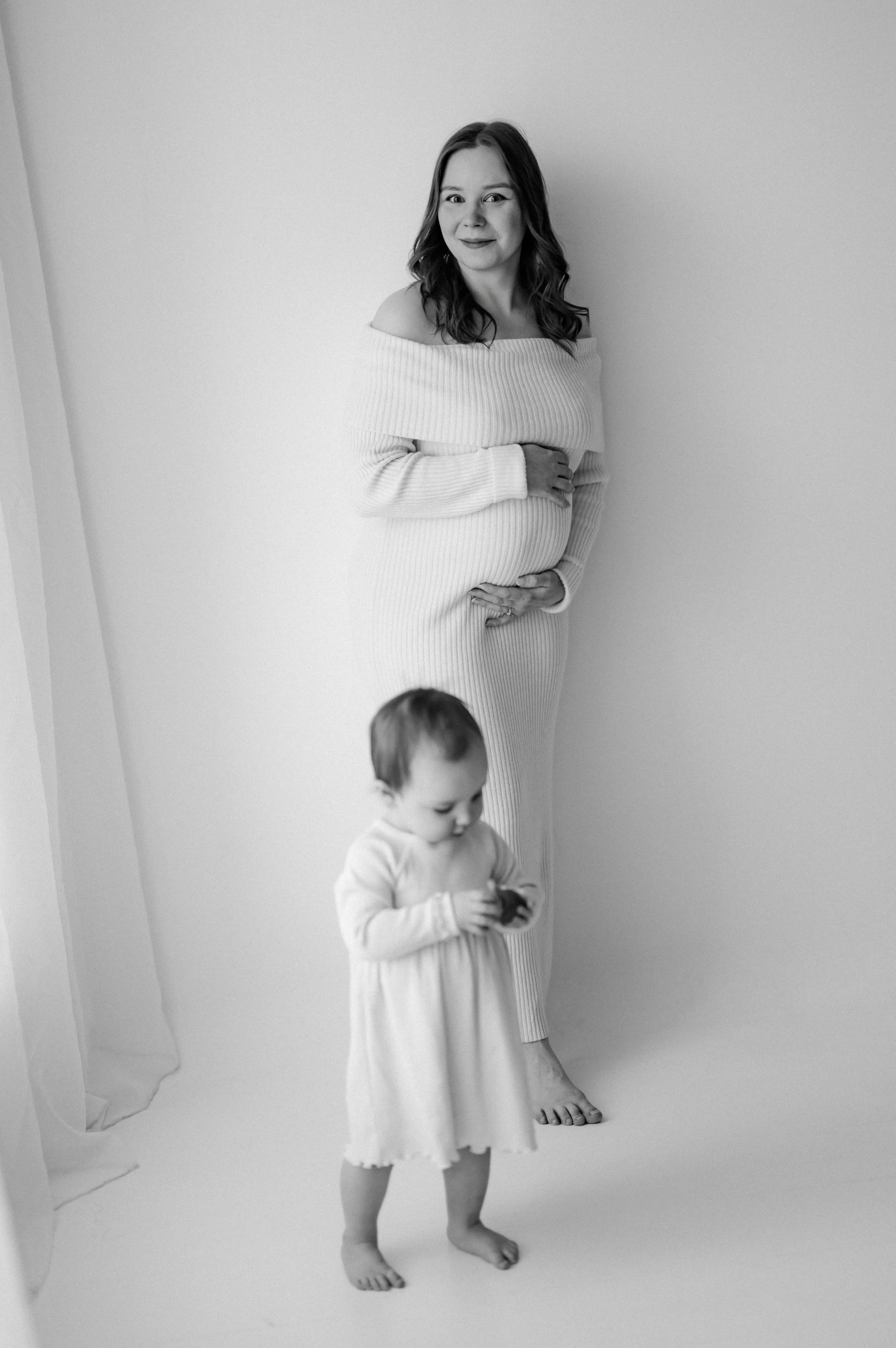 Black and white photo of a pregnant woman standing next to a young girl. The woman is smiling and wearing a knit off-the-shoulder dress, with her hand on her belly. The girl is looking down at an object in her hands, wearing a light dress, and appear