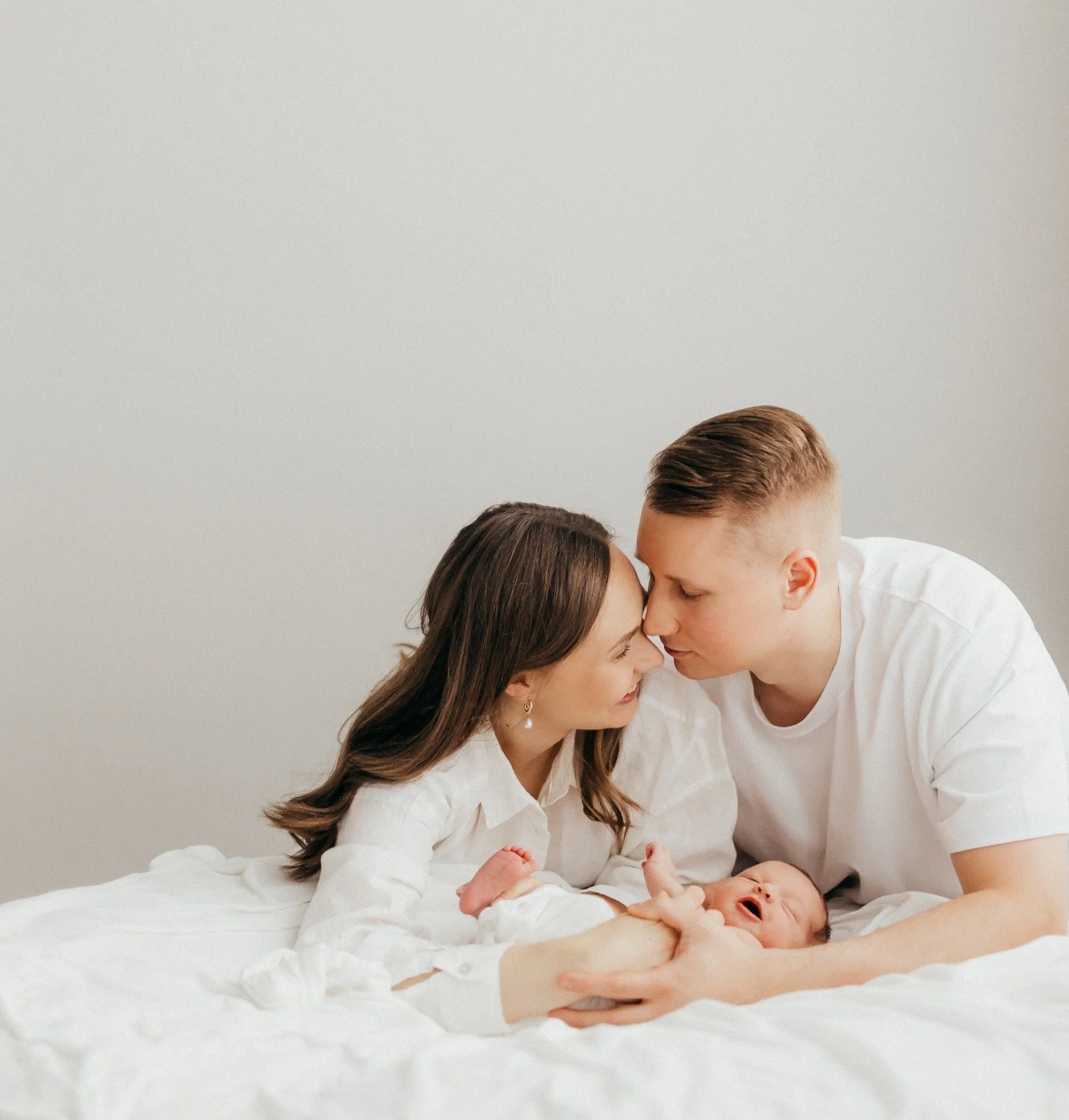A young family with a mother and father holding their crying newborn on a white bed, touching their foreheads together in a loving moment.