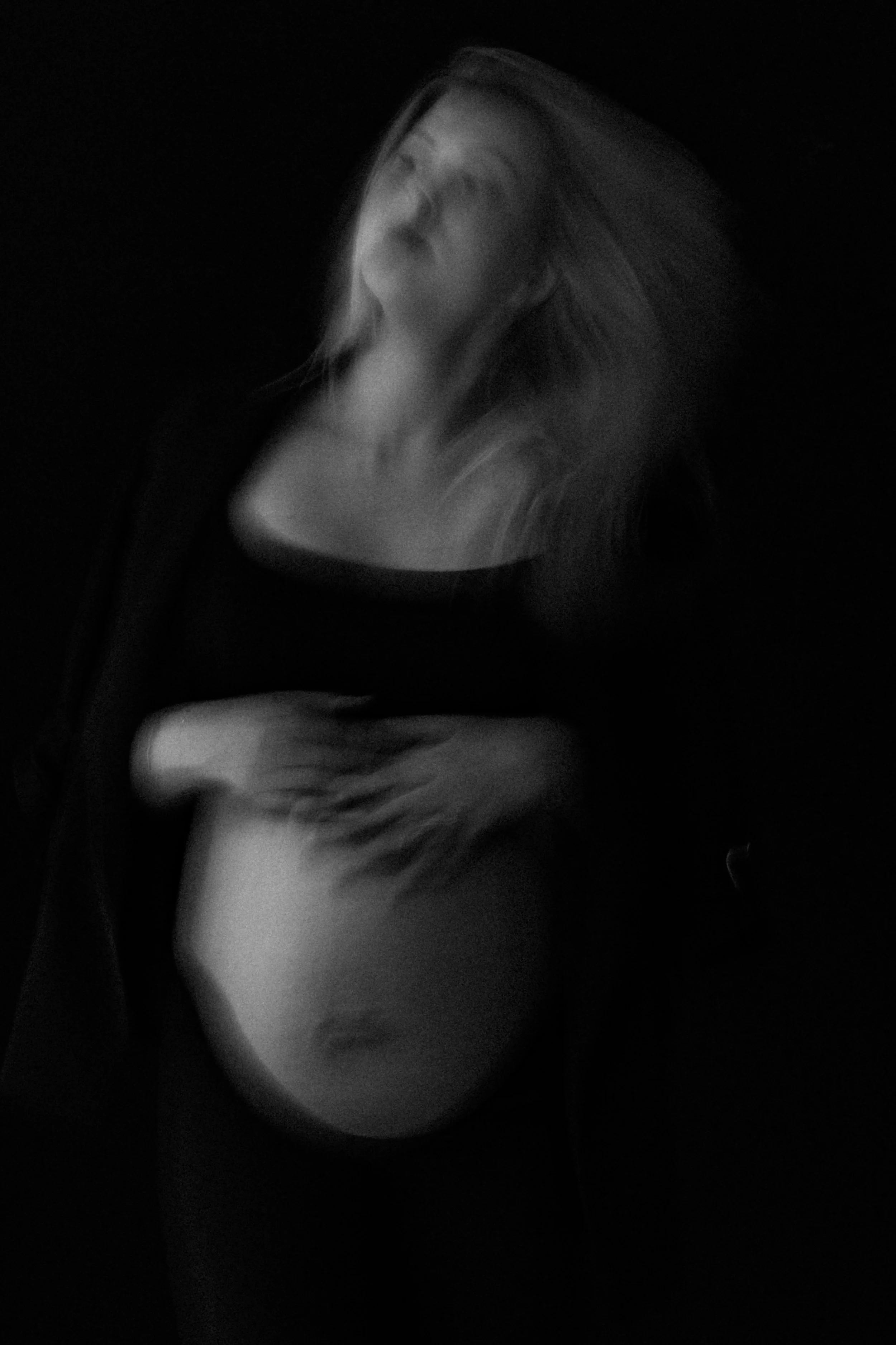 A blurred black and white photograph of a woman with long hair, holding her pregnant belly with one hand and her other hand placed on her chest, in a dark environment.