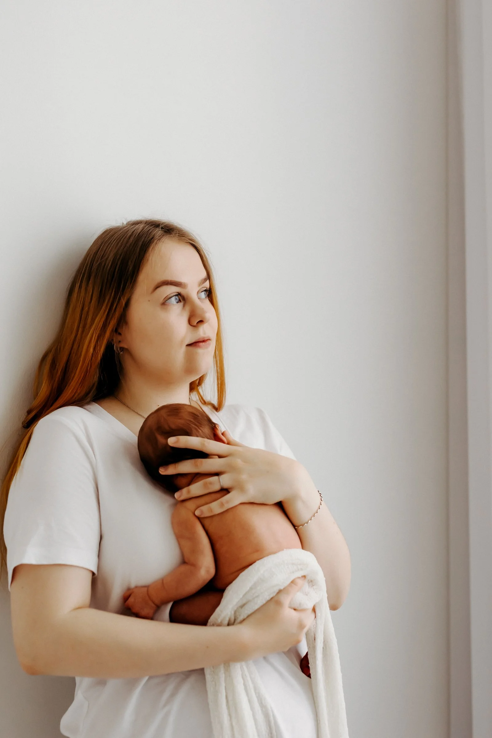 A young woman with red hair sitting against a white wall, holding a newborn baby on her chest, looking off to the side.