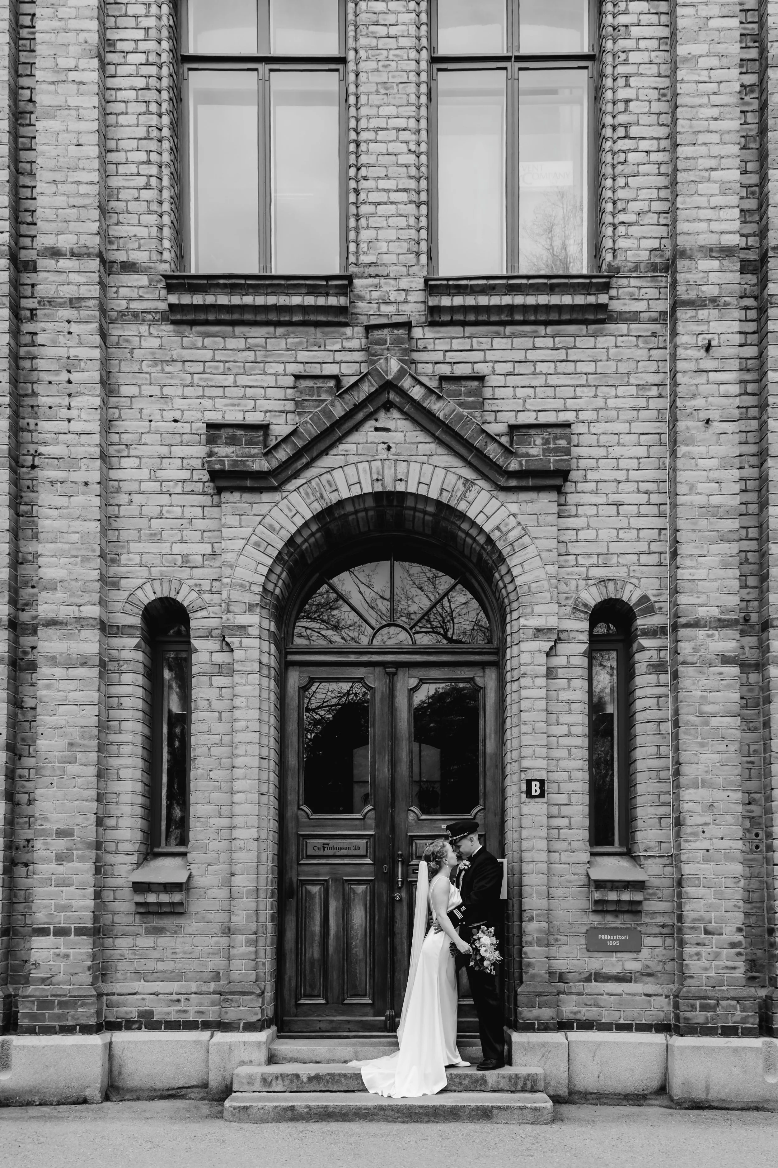A bride and groom in wedding attire sharing a kiss outside a brick building with a large wooden door.
