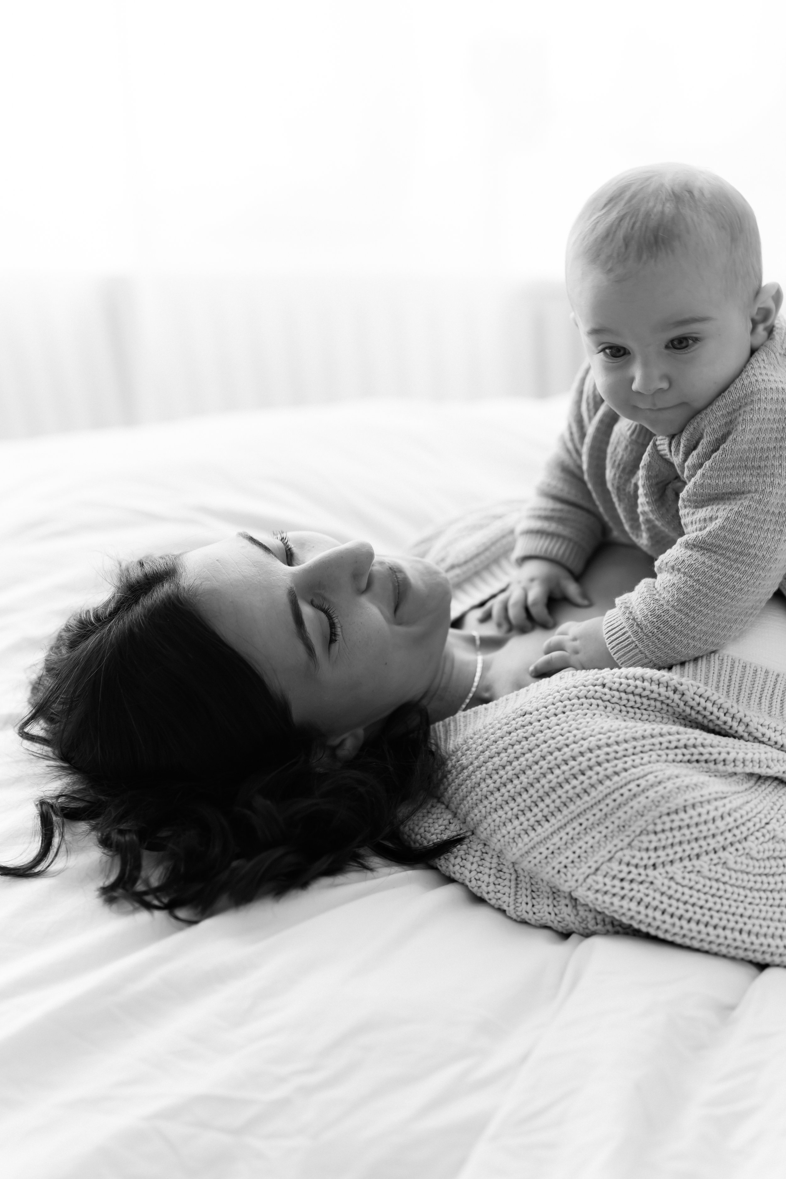 A woman lying on a bed and a young child crawling on her, both smiling and looking at each other.
