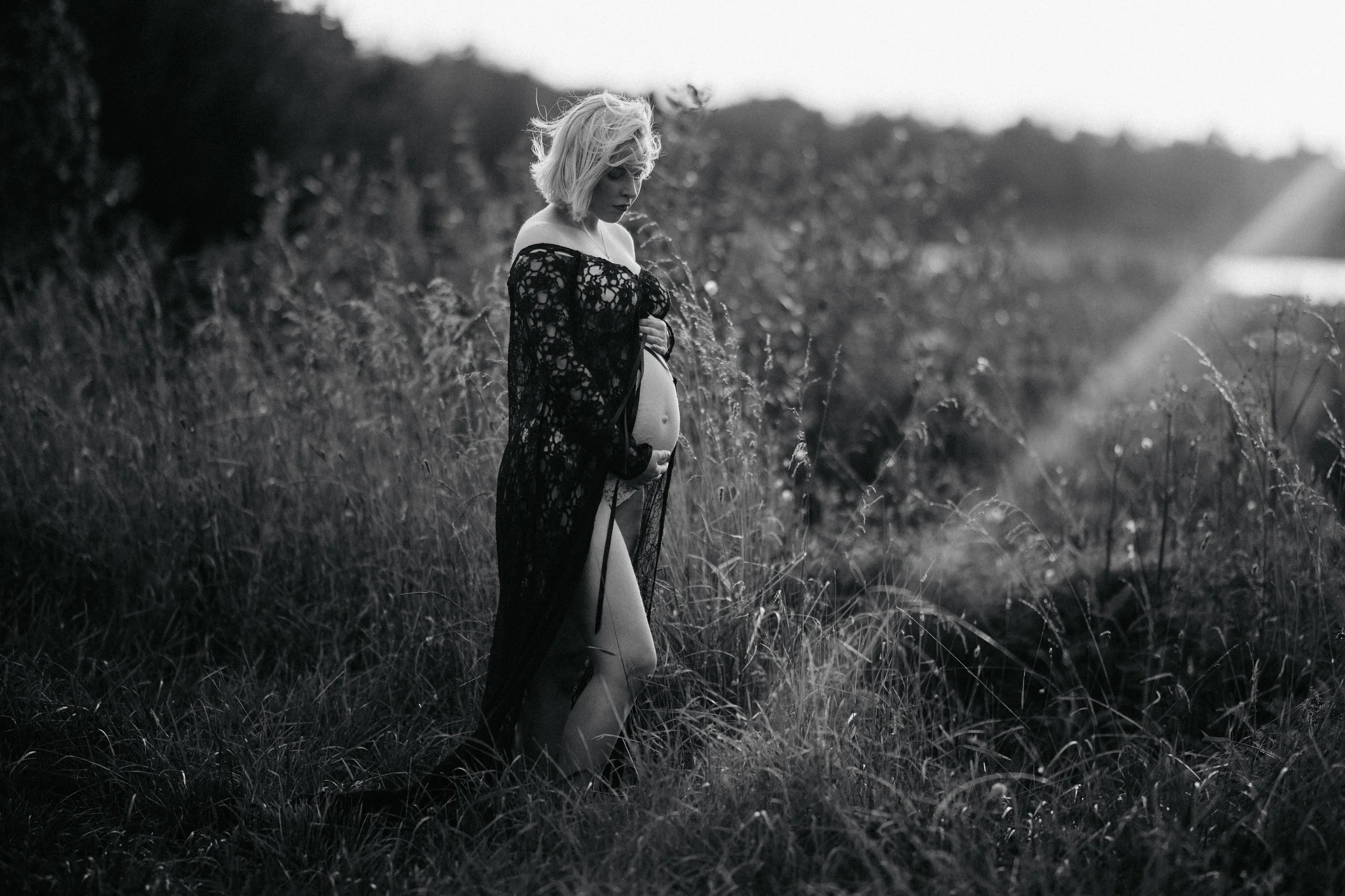 A pregnant woman stands in a grassy field wearing a sheer, lace robe, looking down and touching her pregnant belly.