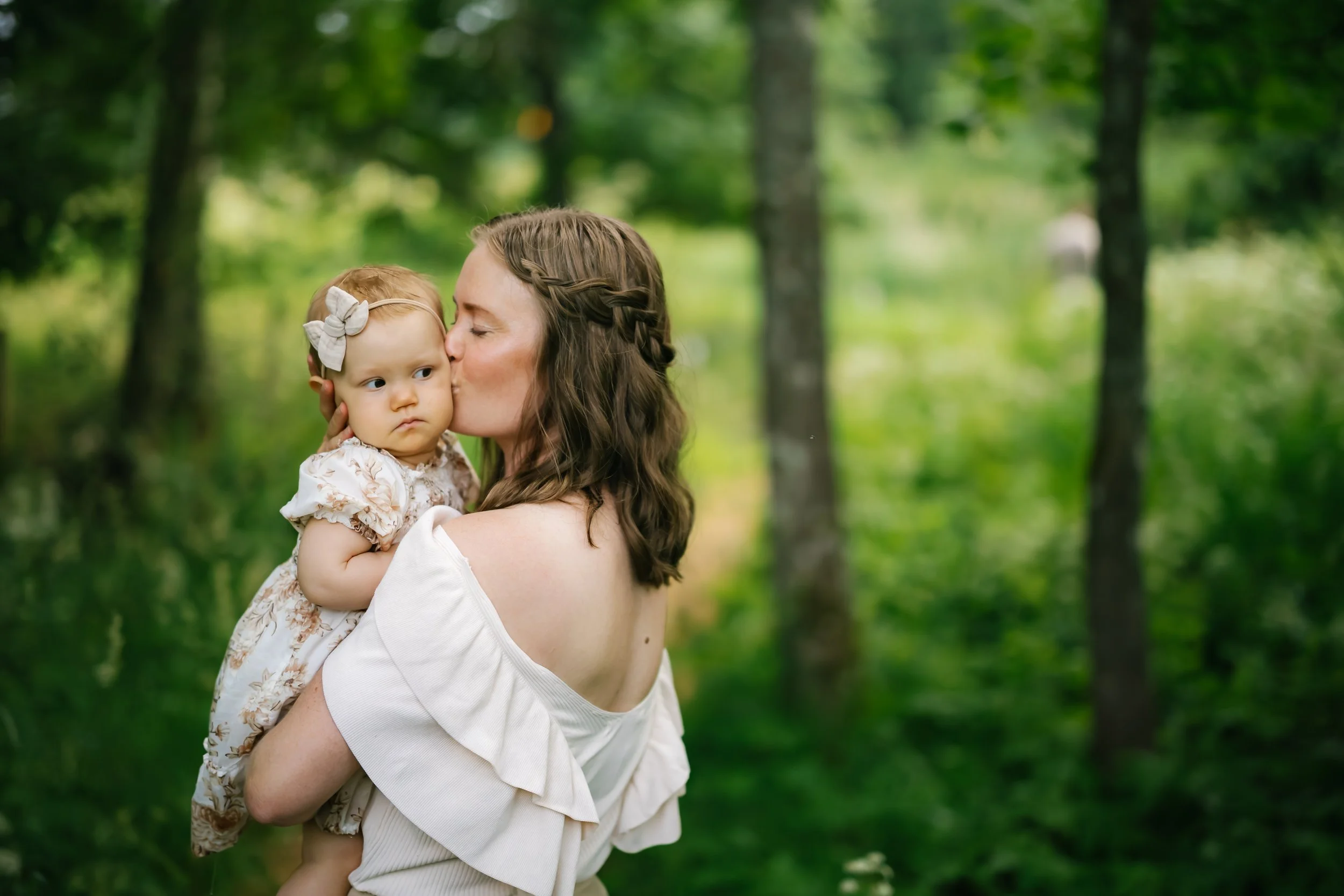 A woman with brown hair, wearing a white off-shoulder top, kissing a young girl with a cream-colored bow headband, in a lush green forest.