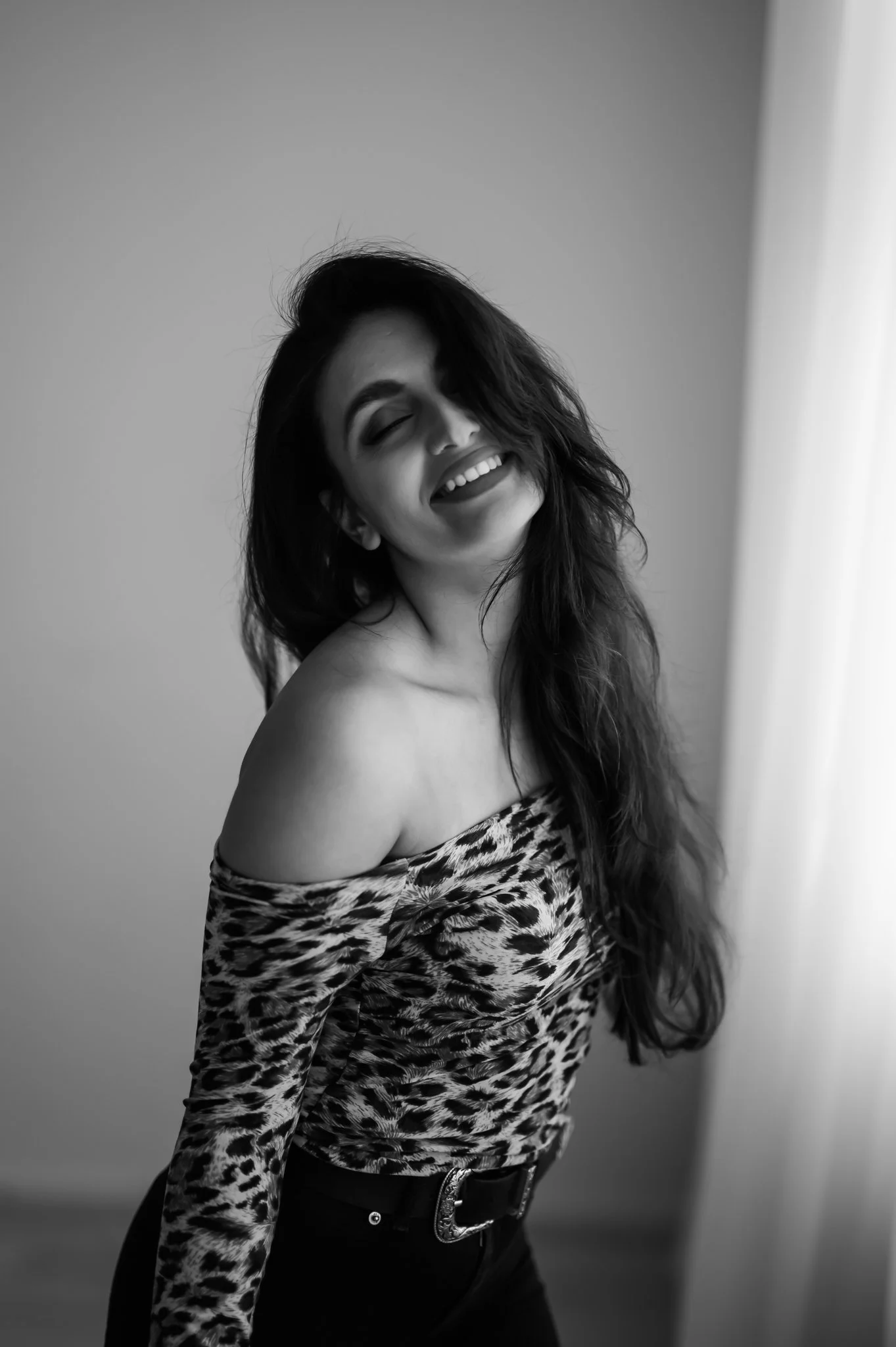 A woman with long hair, smiling with closed eyes, wearing an off-shoulder leopard print top and black pants, standing near a curtain.