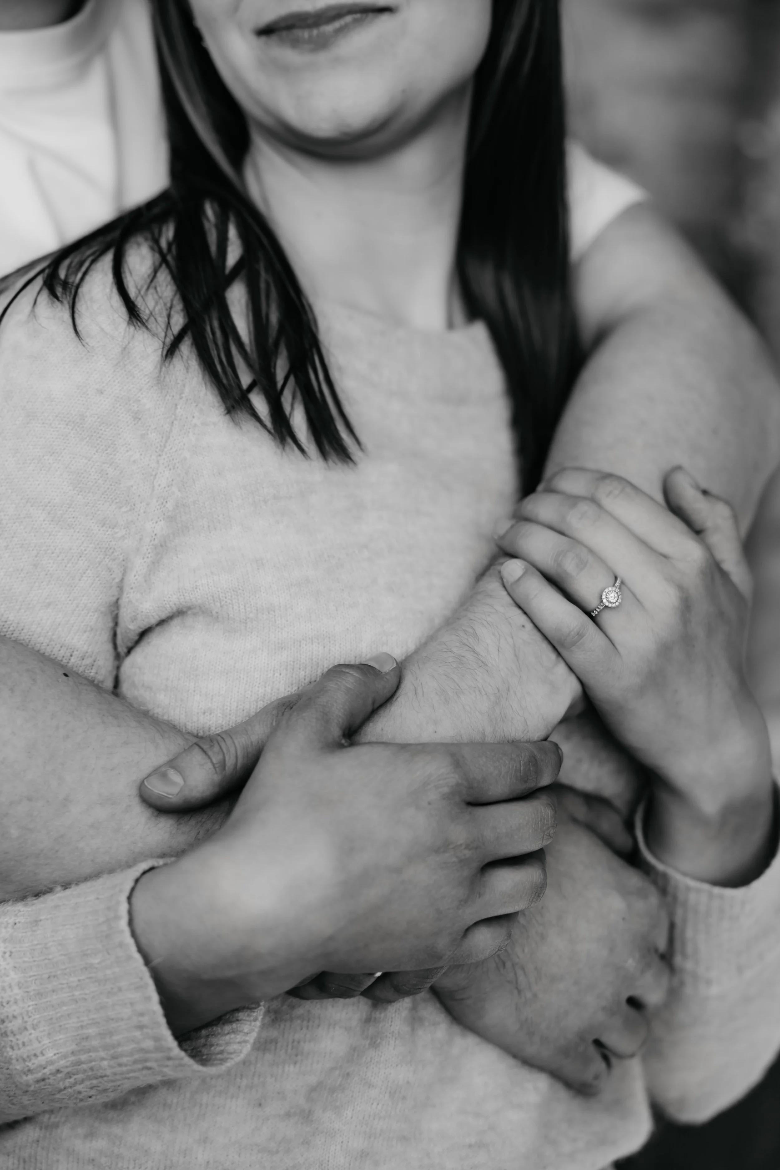 Close-up of a woman with dark hair, wearing a ring, being embraced by a person with light skin, in black and white.