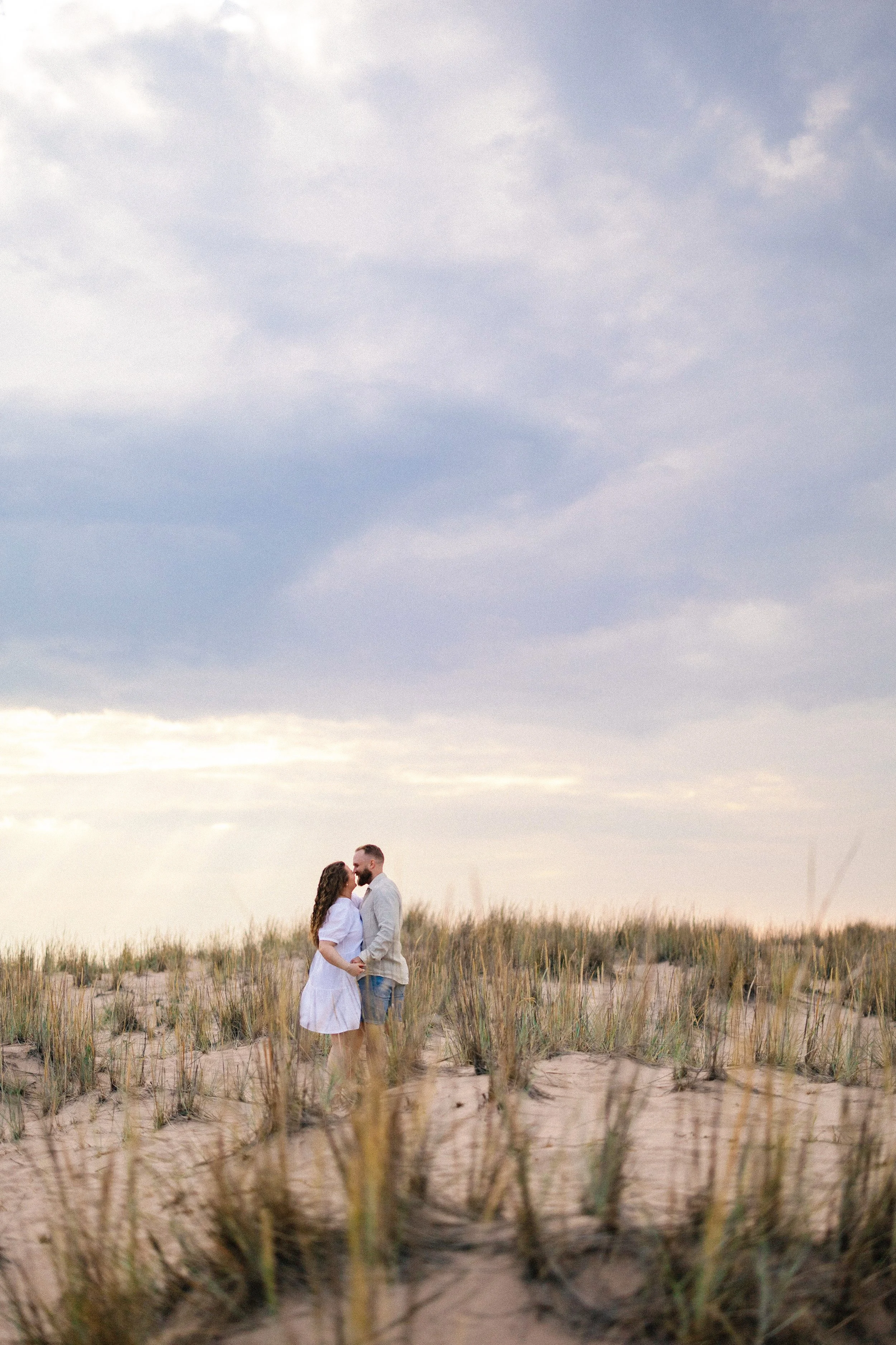 A couple holding hands stands close together on a sandy beach with grassy dunes, under a cloudy sky during sunset.