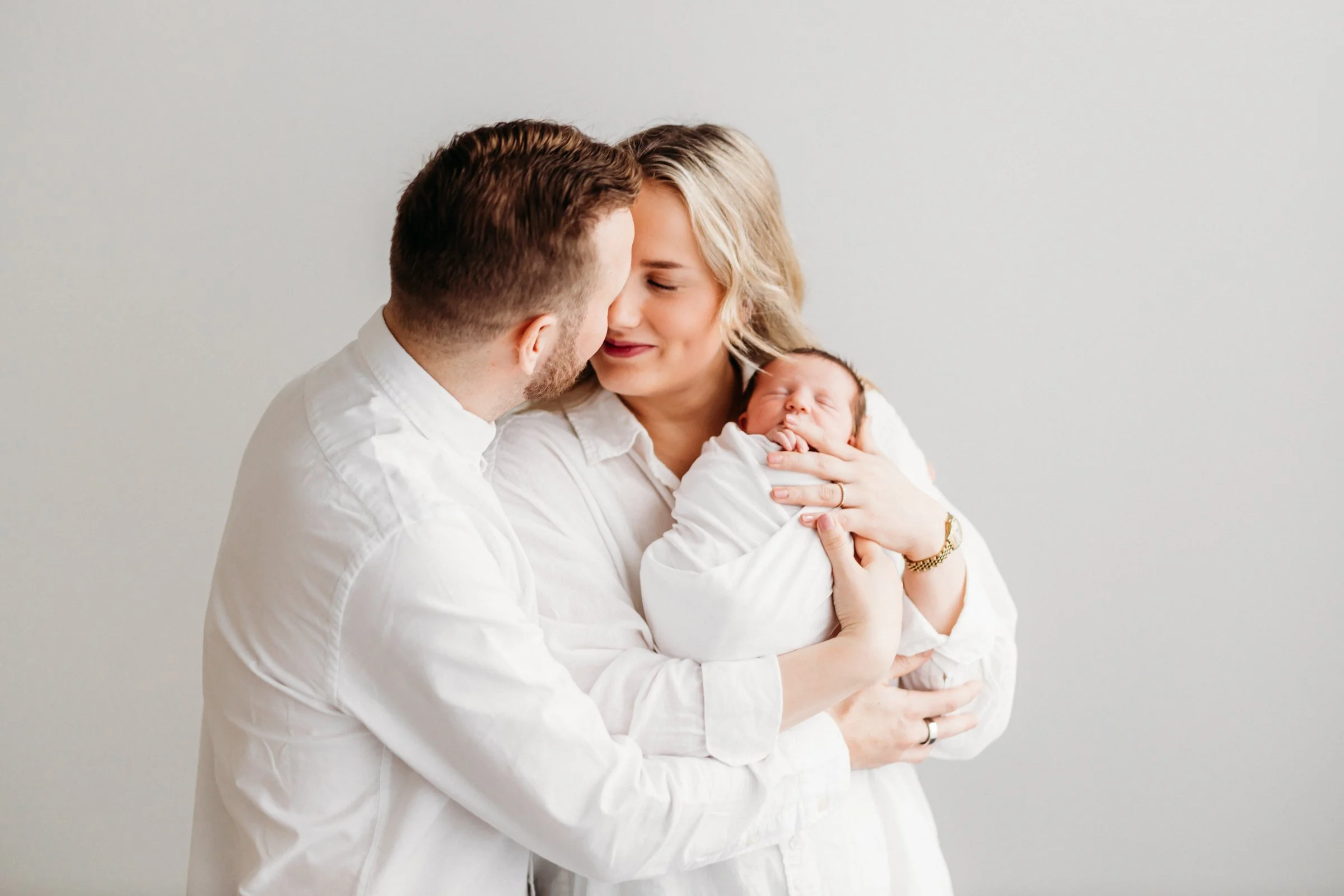 A couple holding a newborn baby and touching foreheads, all dressed in white against a plain light background.