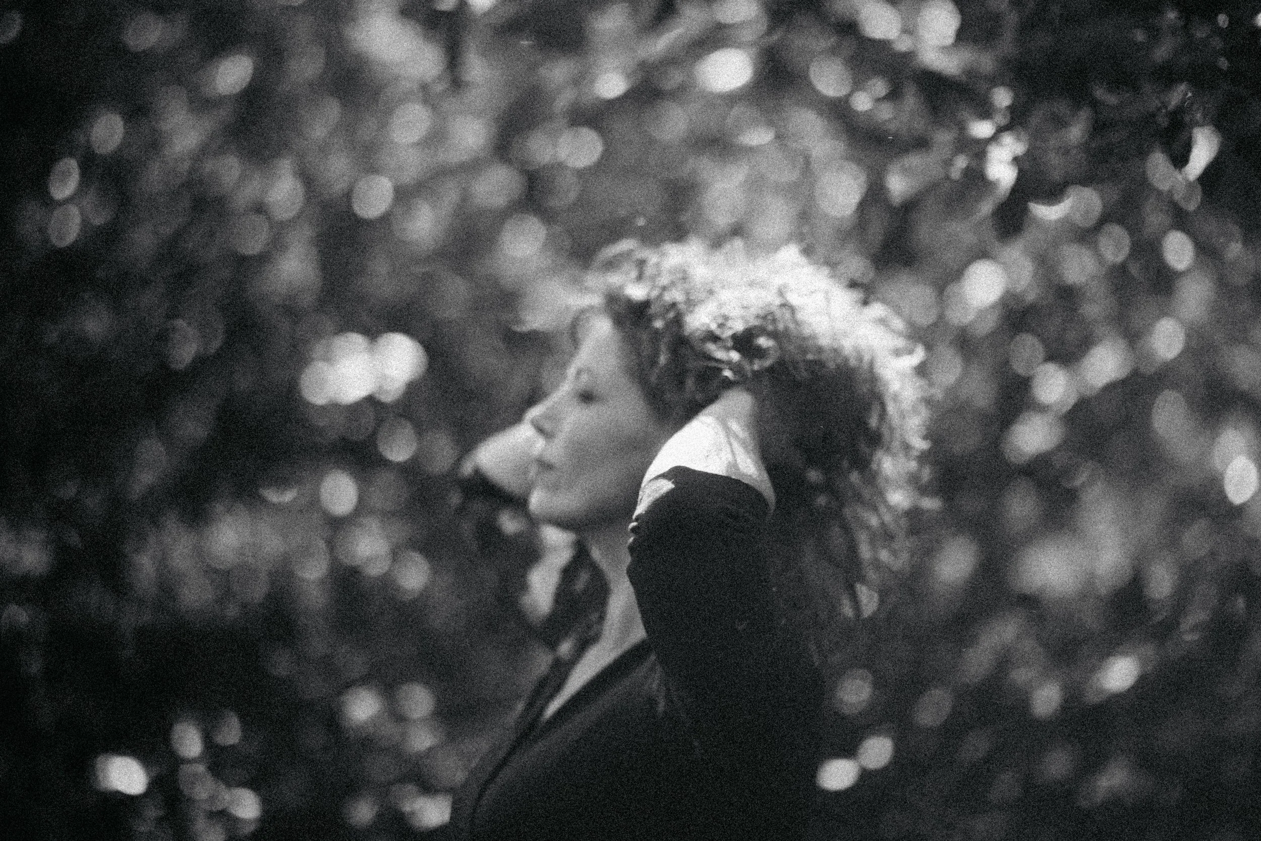 Black and white photo of a woman with curly hair, eyes closed, resting her head on her hand, with sunlight through trees in the background.