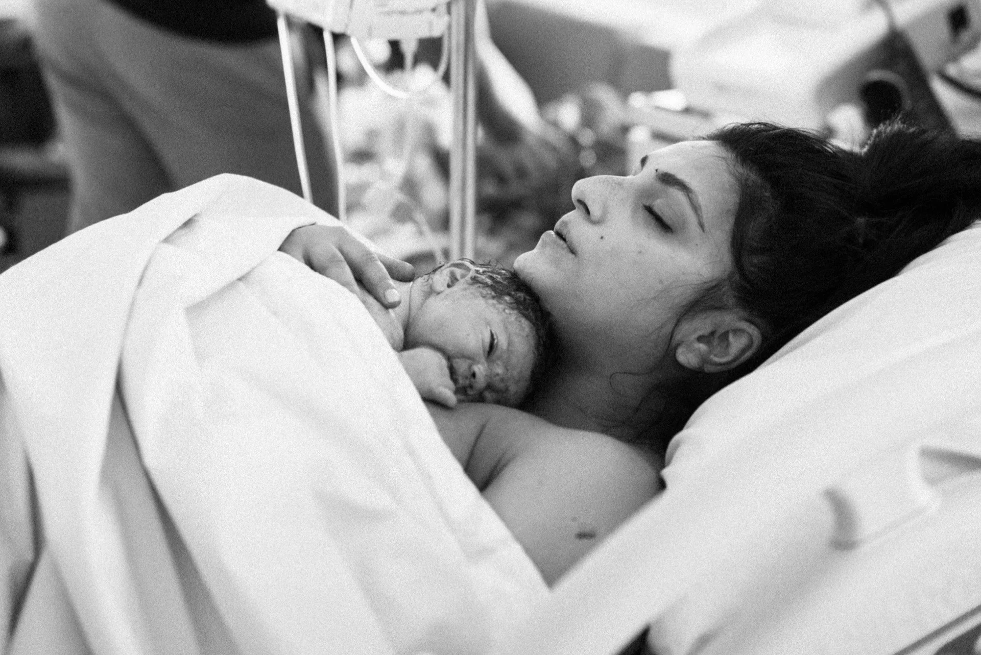 A woman with dark hair lies in a hospital bed, holding a newborn baby close to her chest, both appearing to be resting after childbirth.