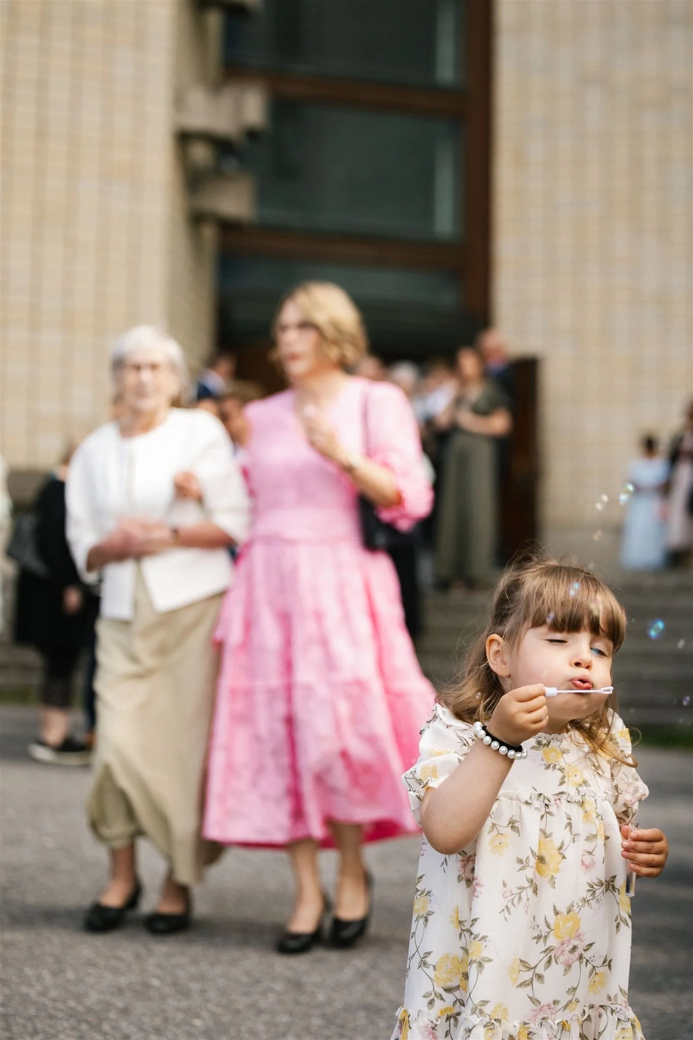 A young girl with brown hair blowing bubbles outdoors, with a pink floral dress and a bracelet, in front of blurred adults including women in pastel and beige outfits, near a brick building.