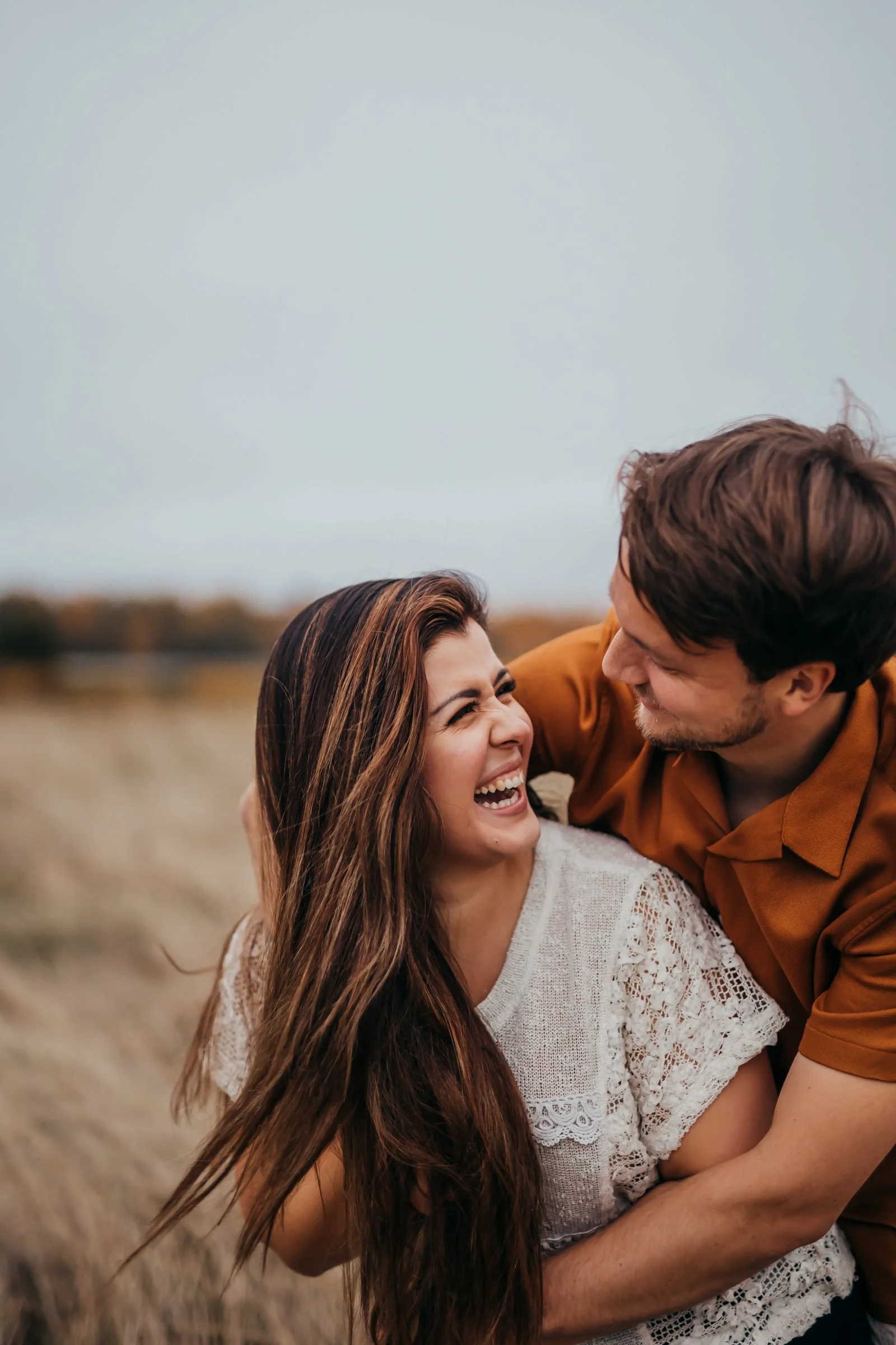 A couple laughing and embracing outdoors in a field with a cloudy sky.