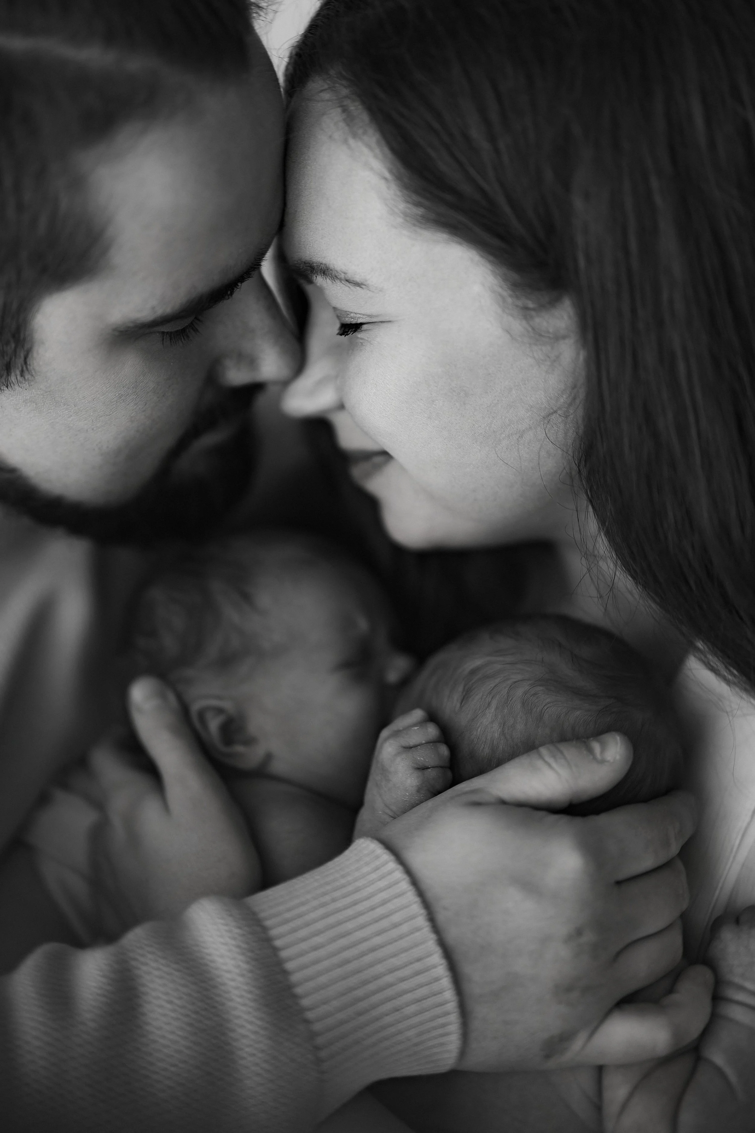 Black and white photo of a couple lovingly holding their newborn babies, with foreheads touching and smiling gently.
