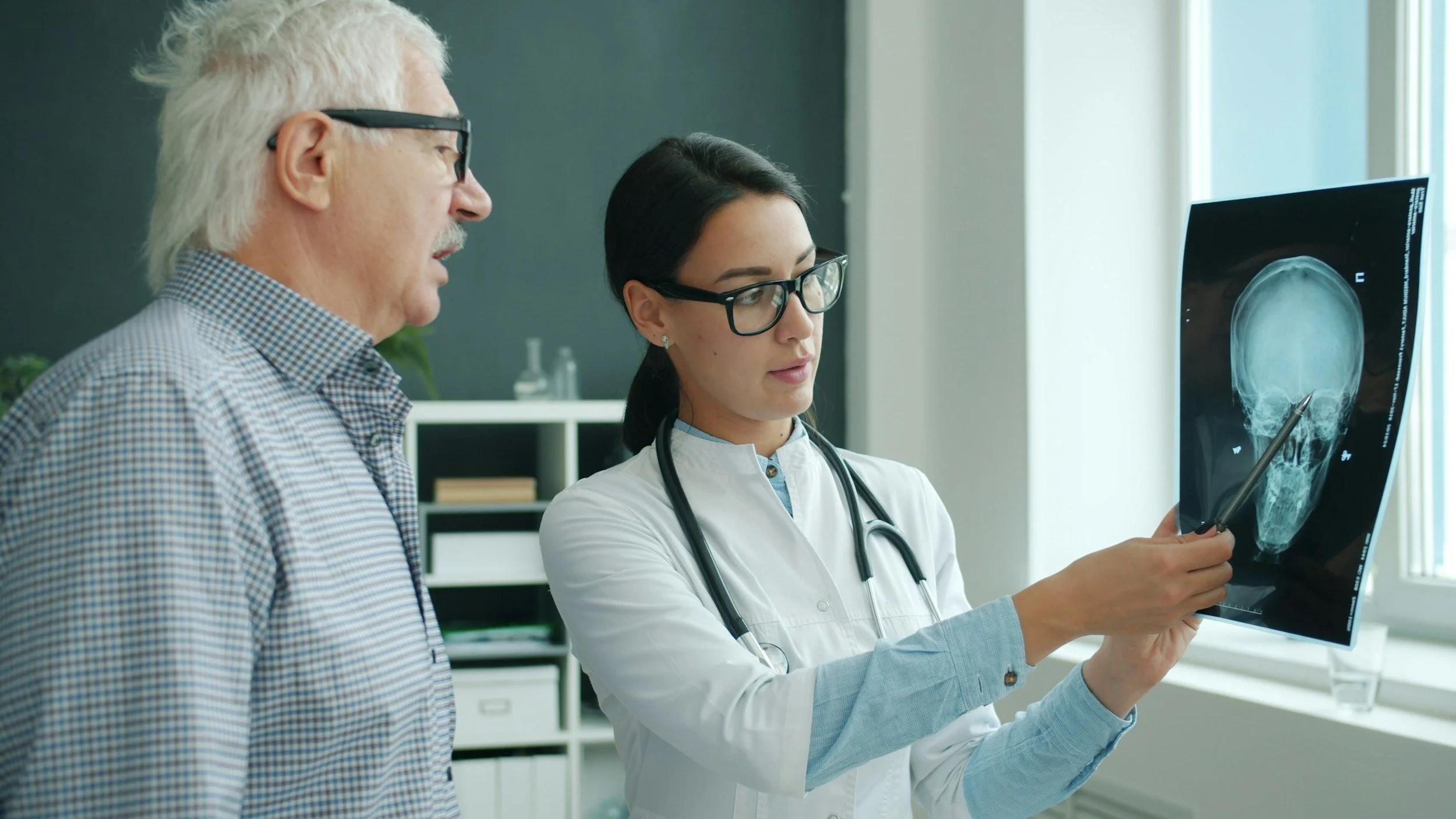 A female doctor showing an X-ray image of a human skull to an older male, possibly a patient, in a medical office.