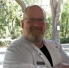 A man with glasses and a beard, smiling outdoors with trees in the background.