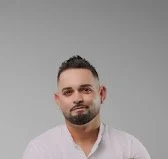 A man with short dark hair and a beard, wearing a light-colored shirt, standing against a plain background.