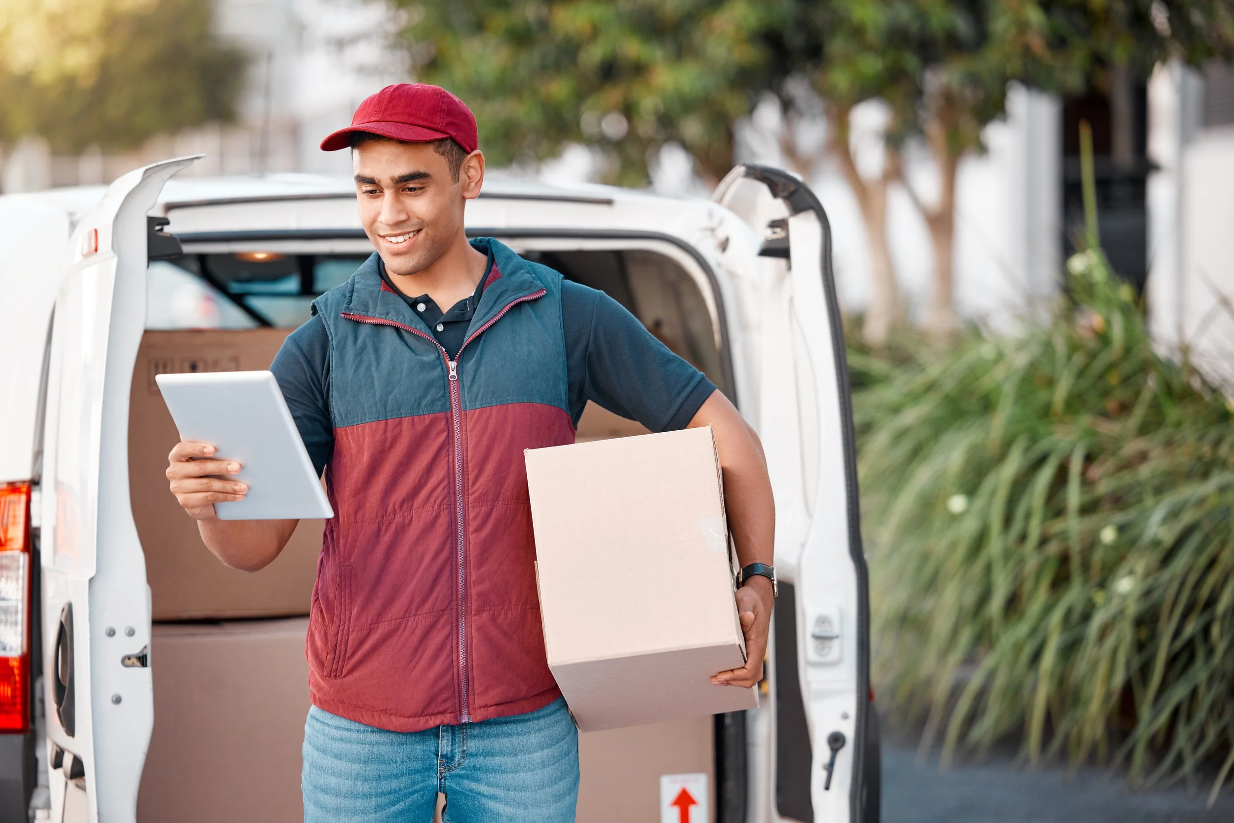 A delivery person holding a cardboard box and looking at a tablet, standing in front of an open delivery van.