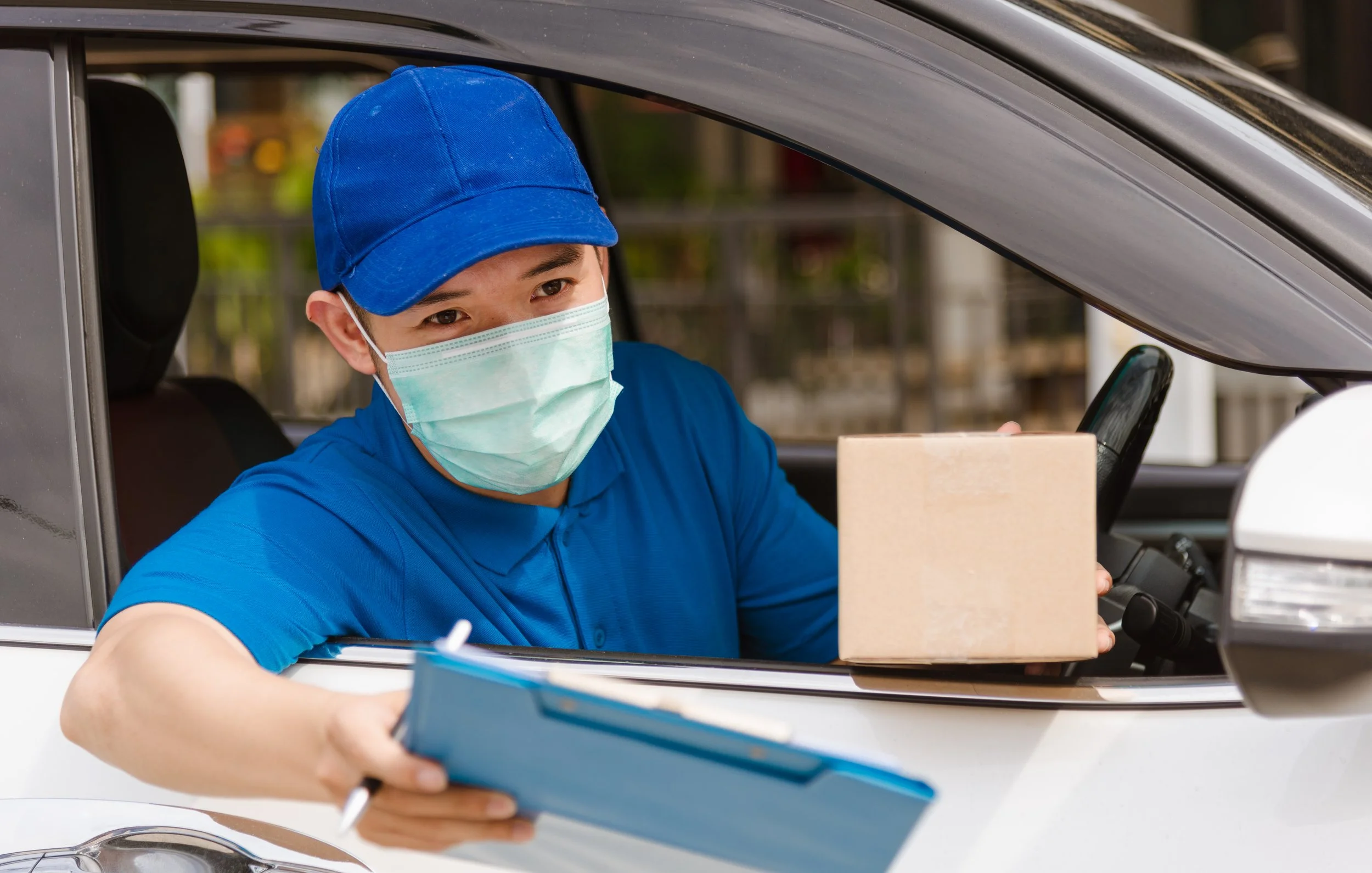 Delivery person wearing a blue uniform, cap, and face mask handing over a package from the vehicle window.
