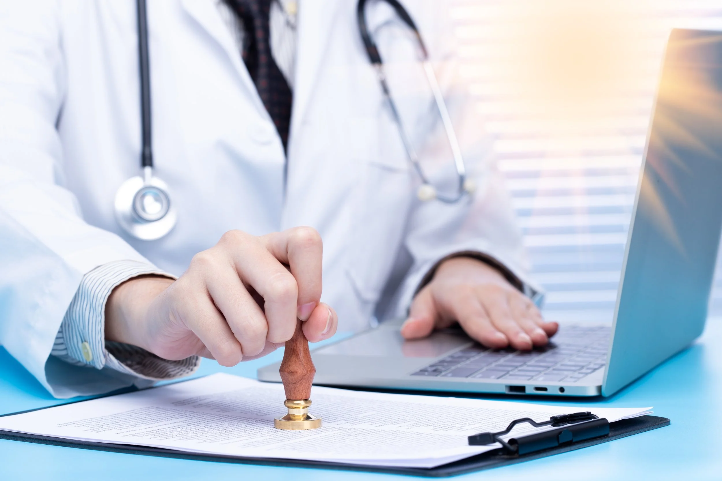 A doctor in a white coat stamps a document with a wooden stamp while working on a laptop at a desk