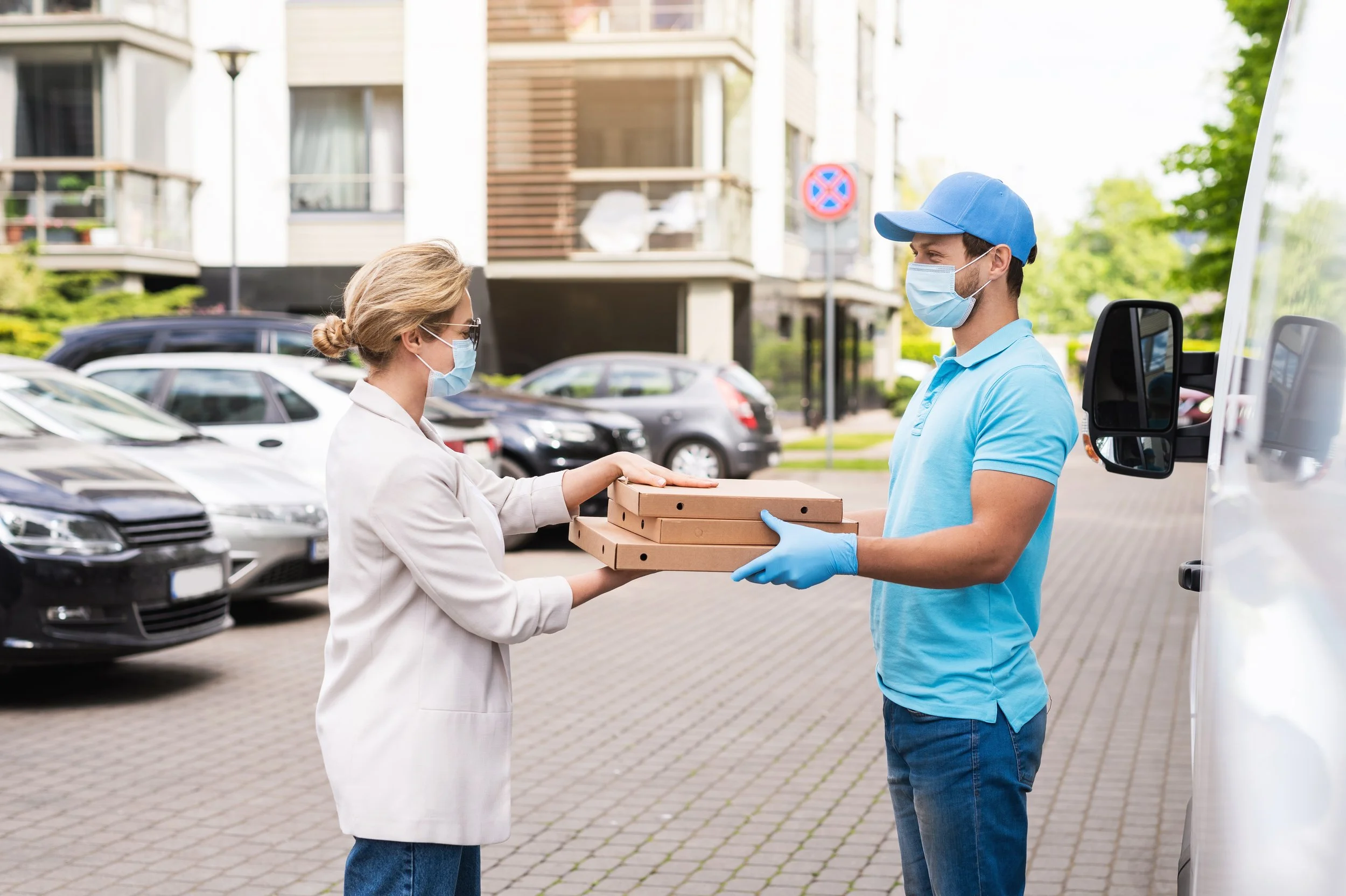 A woman in a light-colored blazer and sunglasses receiving pizza boxes from a delivery person in a light blue polo shirt, cap, gloves, and face mask, outside a residential building. Both are wearing masks.