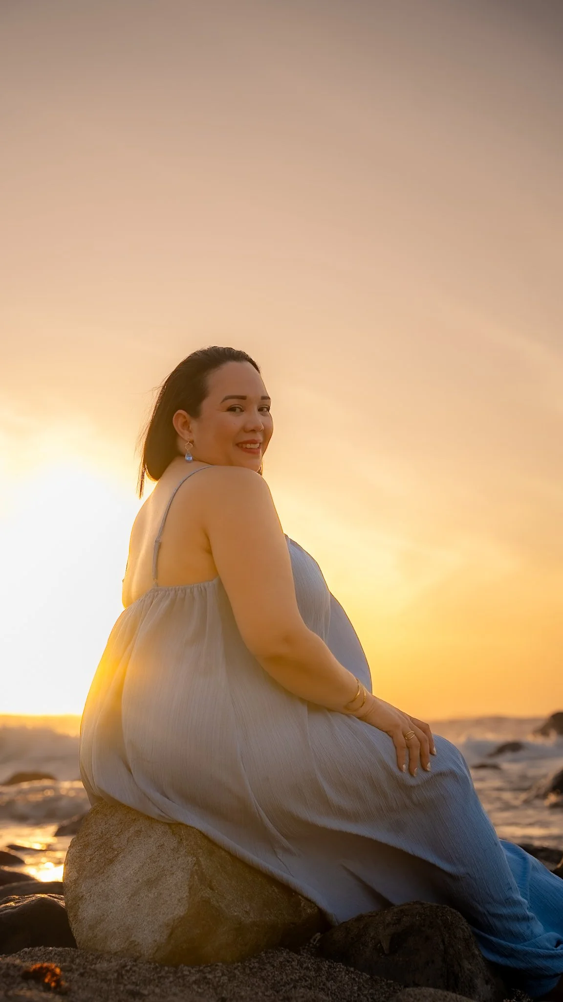 A woman sitting on a rock at the Aruba beach during sunset, smiling and looking over her shoulder.