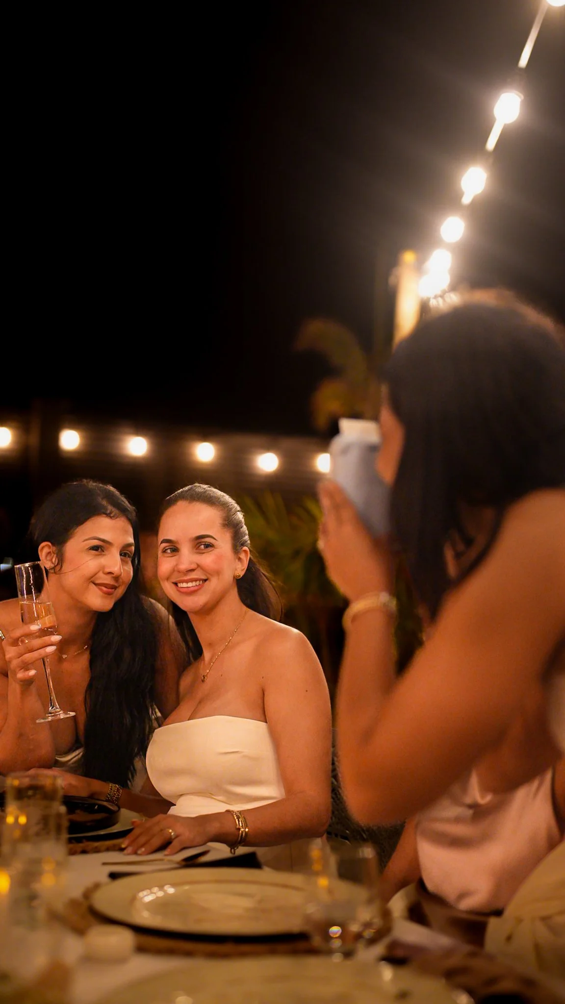 Two women smiling at a dinner party, one holding a champagne glass, with a photographer taking their picture, under string lights at night.